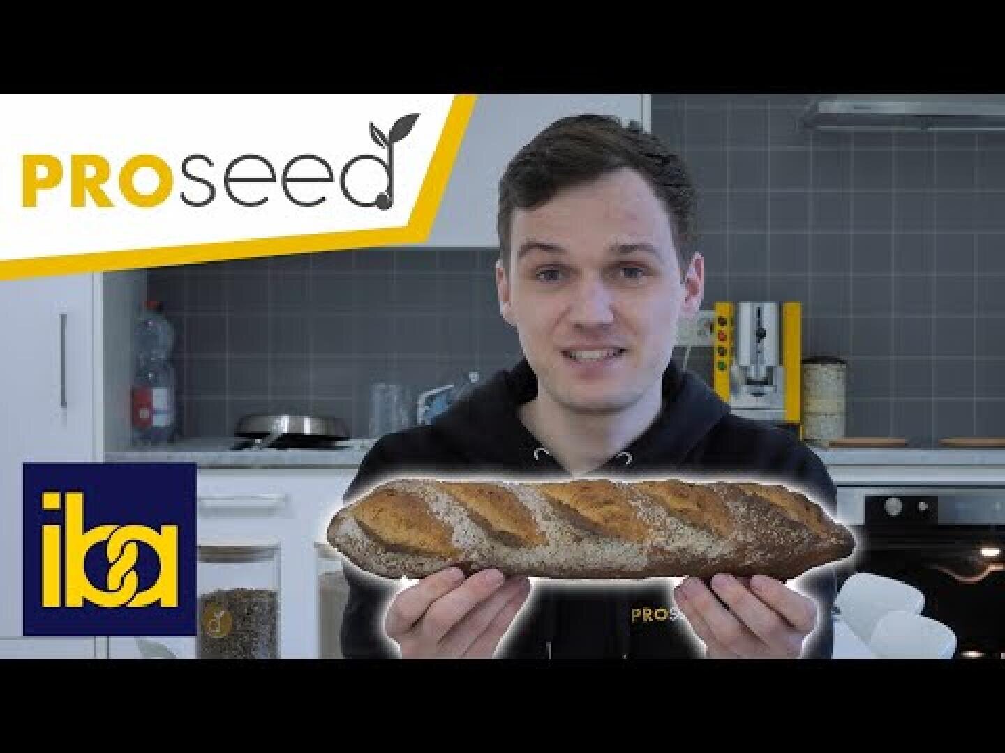 Eine Person in einer Küche hält einen Laib Brot. Das Bild zeigt das PROseed-Logo oben links und das iba-Logo unten links. Im Hintergrund sind Küchengeräte und Behälter zu sehen.