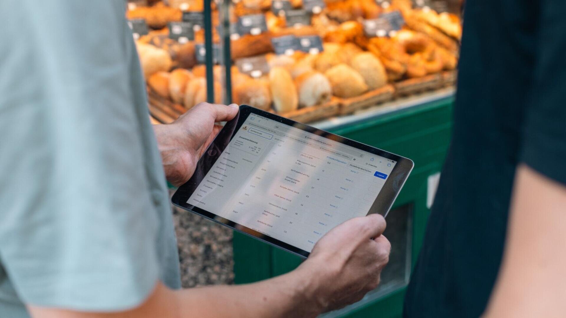 Two people stand in front of a bakery display with pastries, one holding a tablet showing a spreadsheet or digital interface.