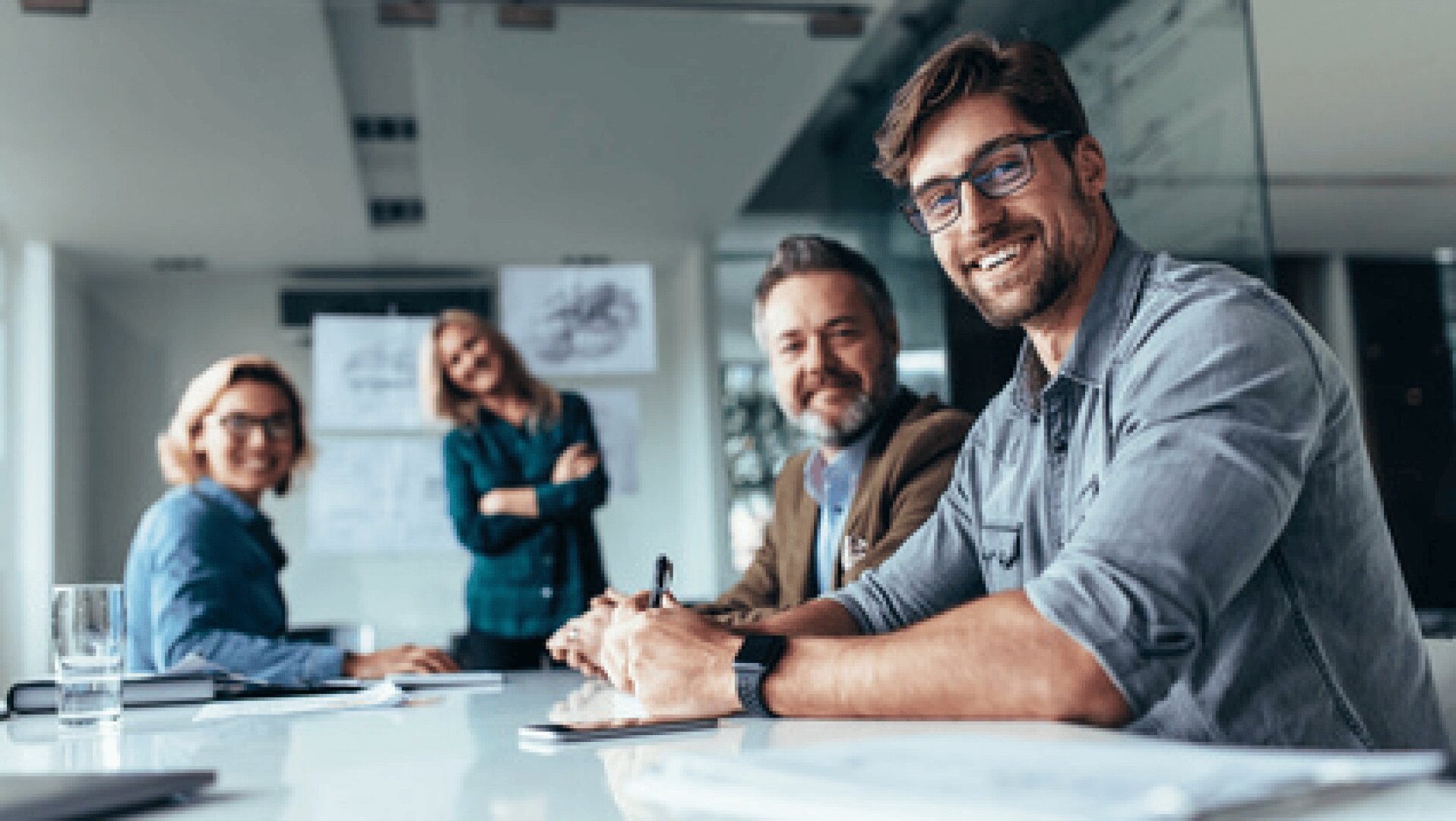 Vier Personen sitzen und stehen um einen Konferenztisch in einem modernen Büro und lächeln in die Kamera. Auf dem Tisch liegen Papiere und ein Glas Wasser, und an der Wand im Hintergrund sind Tabellen zu sehen.
