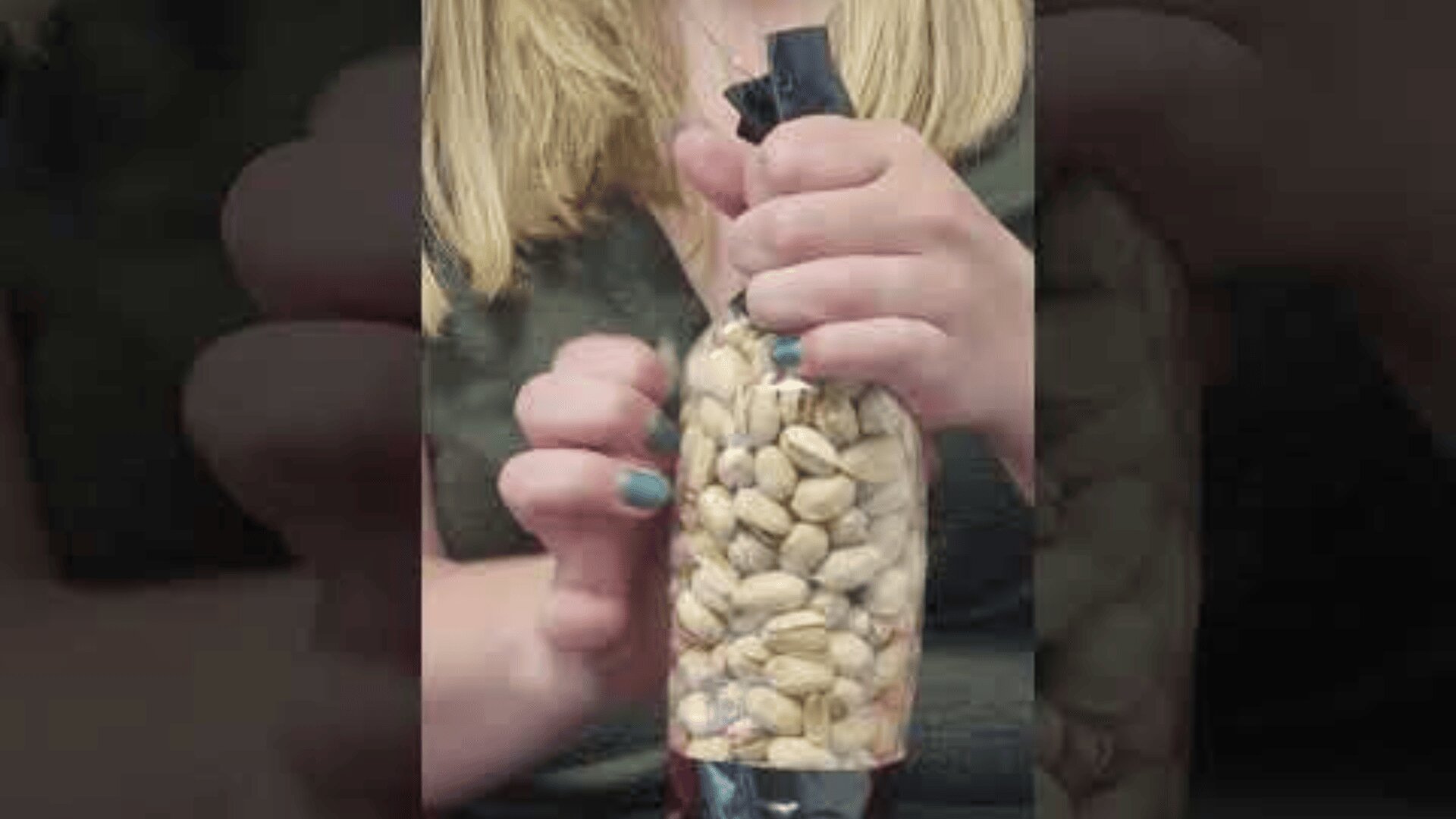 A person with blond hair is holding a plastic bottle filled with pistachios. The bottle has a black lid and the person is wearing blue nail polish.
