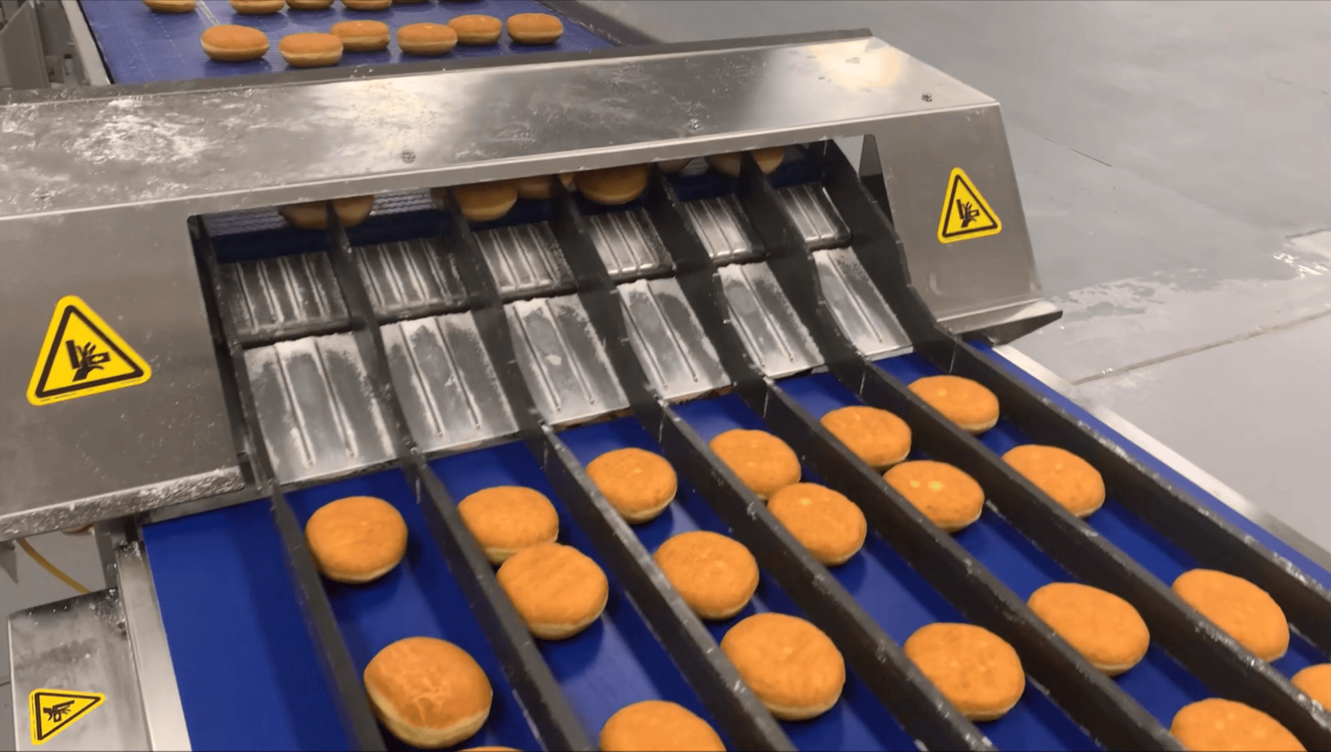 Rows of round donuts move along conveyor belts through a stainless steel machine in a factory setting. Warning signs with an electrical hazard symbol are visible on the machine.