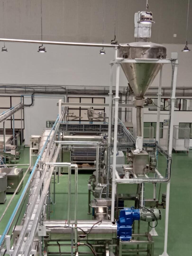 A large industrial food processing machine with metal pipes, tanks, and a funnel-like hopper stands on a green floor in a clean, modern factory setting with overhead lighting.
