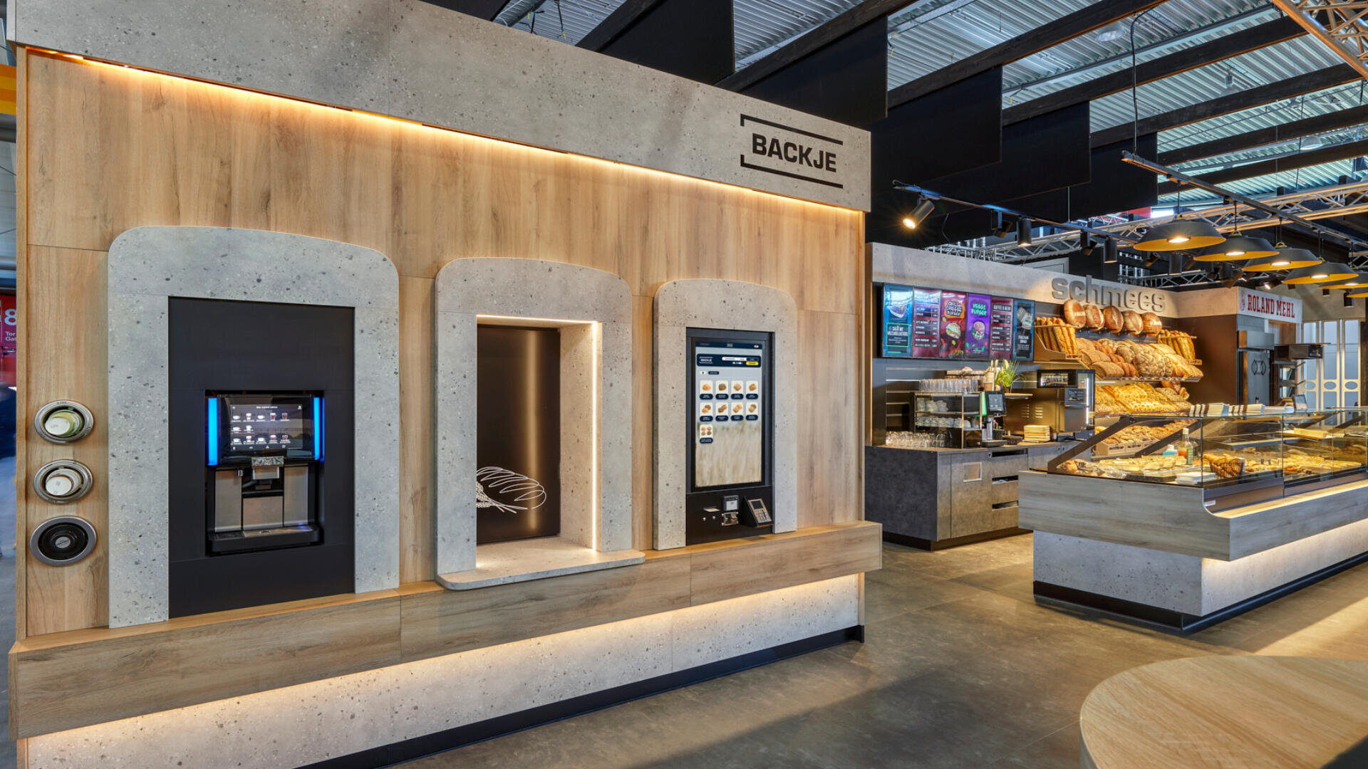 Modernes Bäckereiinterieur mit Selbstbedienungskiosken an einer Holzwand, Brotvitrinen und Regalen, die mit verschiedenen Backwaren gefüllt sind. Im Hintergrund sind helle Beleuchtung und bunte Menüschilder zu sehen.