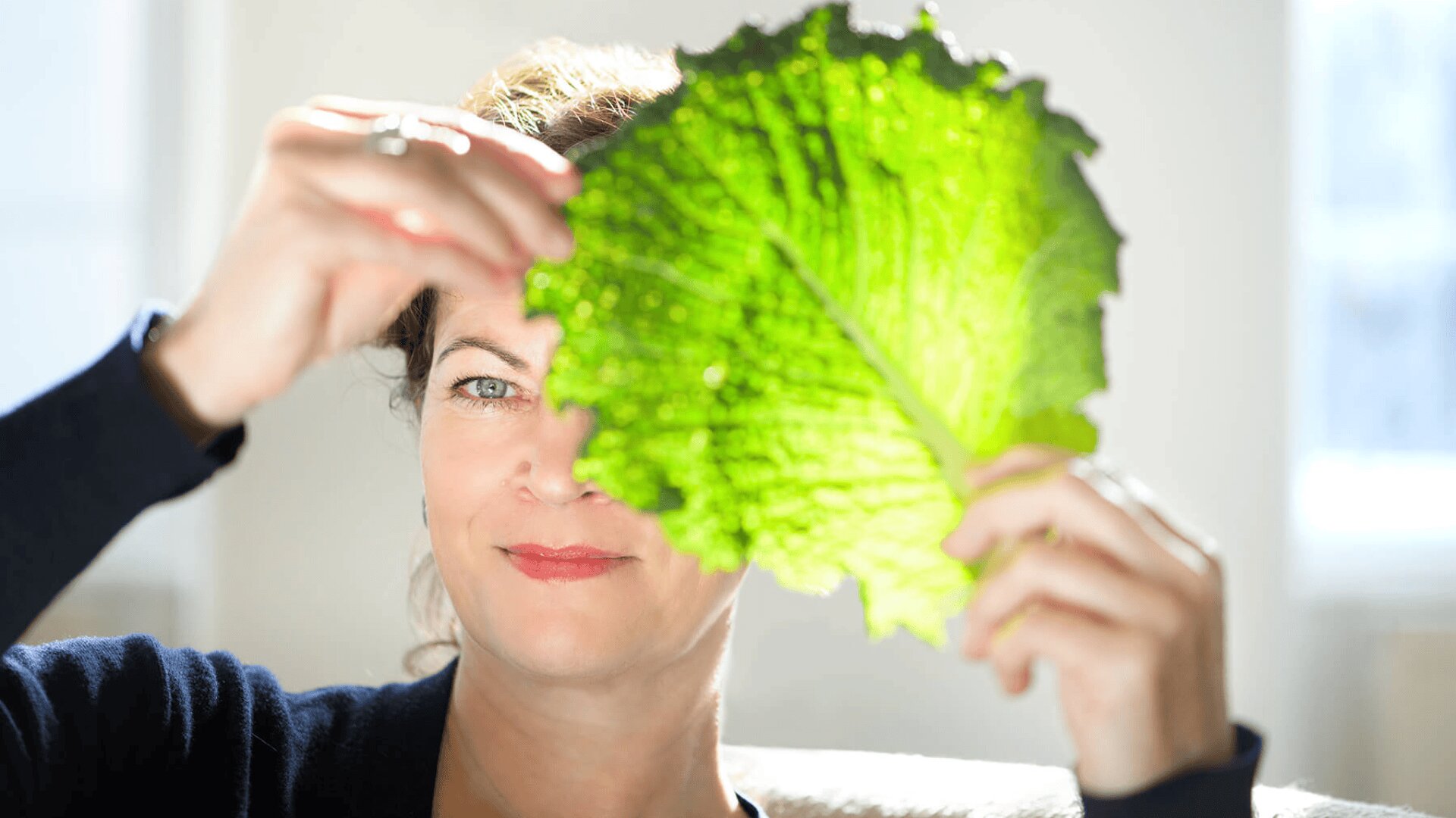 Eine Frau hält ein großes grünes Blatt vor ihr Gesicht und lächelt, während das Sonnenlicht durch das Blatt scheint und seine Struktur und Adern hervorhebt.