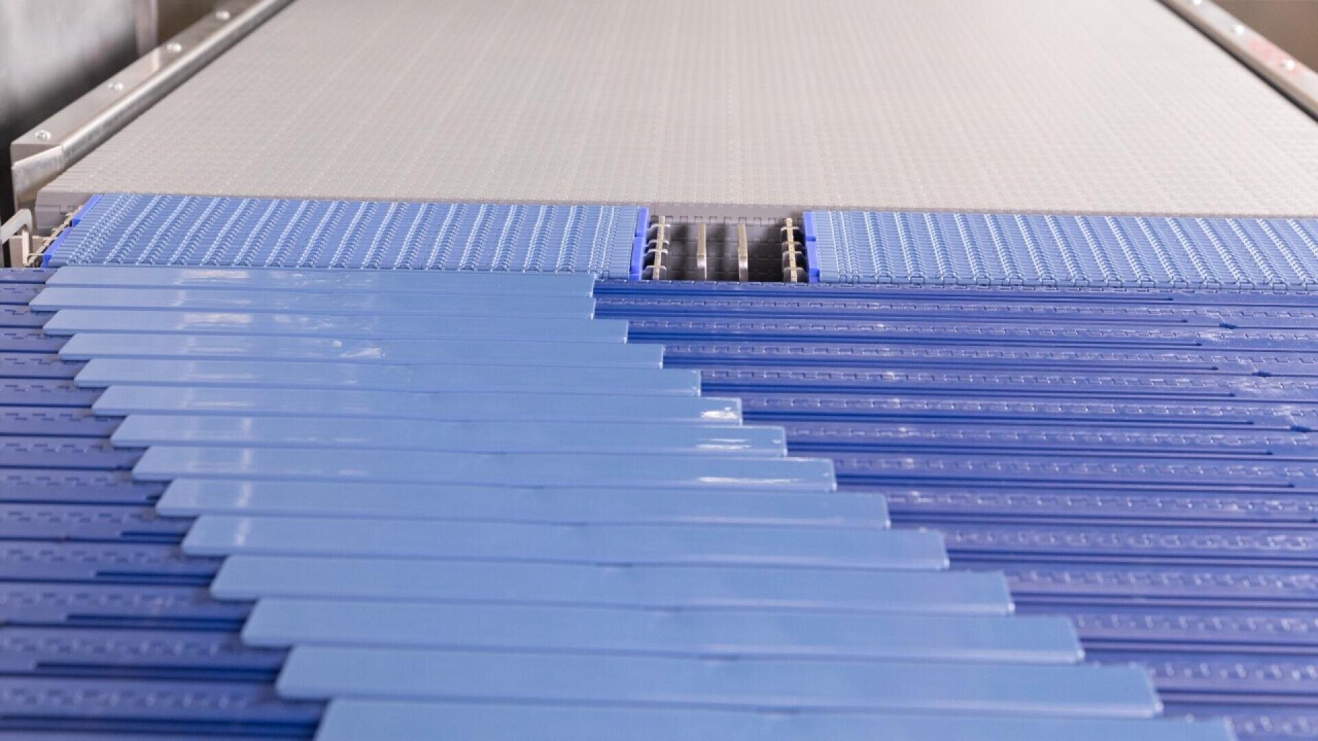 Nahaufnahme eines Fördersystems mit hellblauen Kunststofflamellen, die im Zickzackmuster angeordnet sind und zu einem breiteren Förderband im Hintergrund führen, das wahrscheinlich in Industrie- oder Fertigungsprozessen verwendet wird.