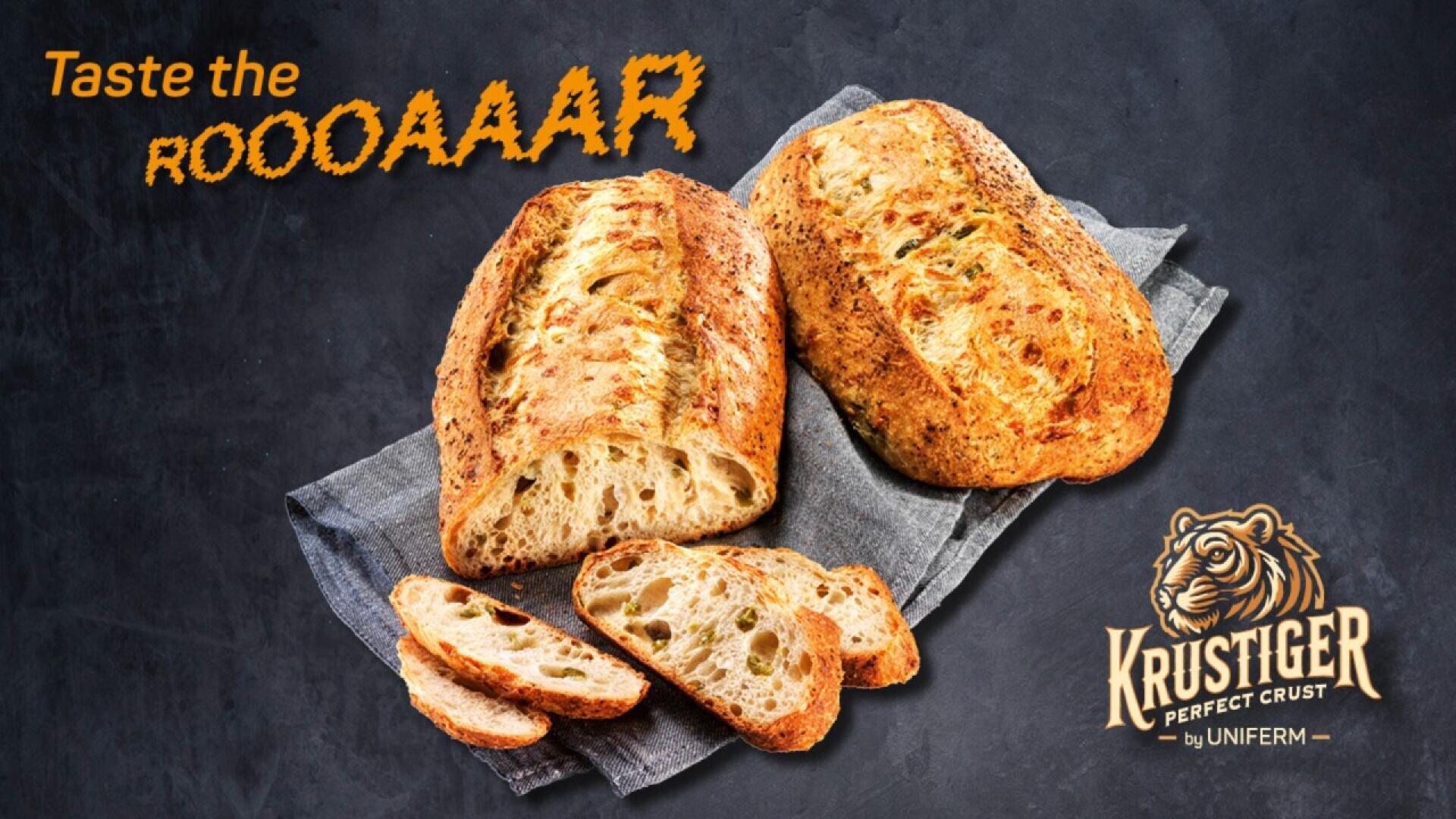 Two loaves of rustic bread, one partially sliced, on a cloth napkin over a dark background. Text reads, Taste the ROOOAAAR with a tiger logo and Krustiger Perfect Crust by UNIFERM in the bottom right corner.
