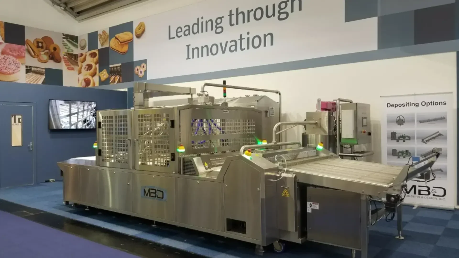 A large stainless steel industrial food processing machine is displayed at an exhibition booth with a sign that reads Leading through Innovation and images of baked goods on the wall.