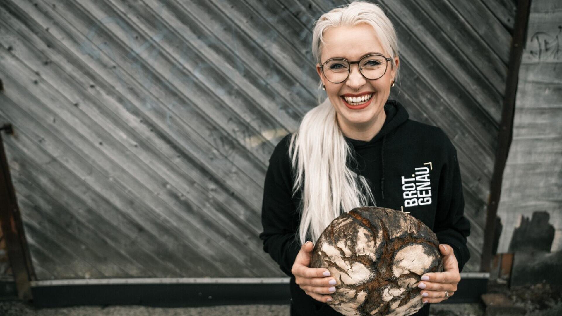 A smiling person with long, light hair and glasses holds a round loaf of rustic bread while wearing a black hoodie with “BROT GENAU” printed on it, standing in front of a weathered wooden wall.