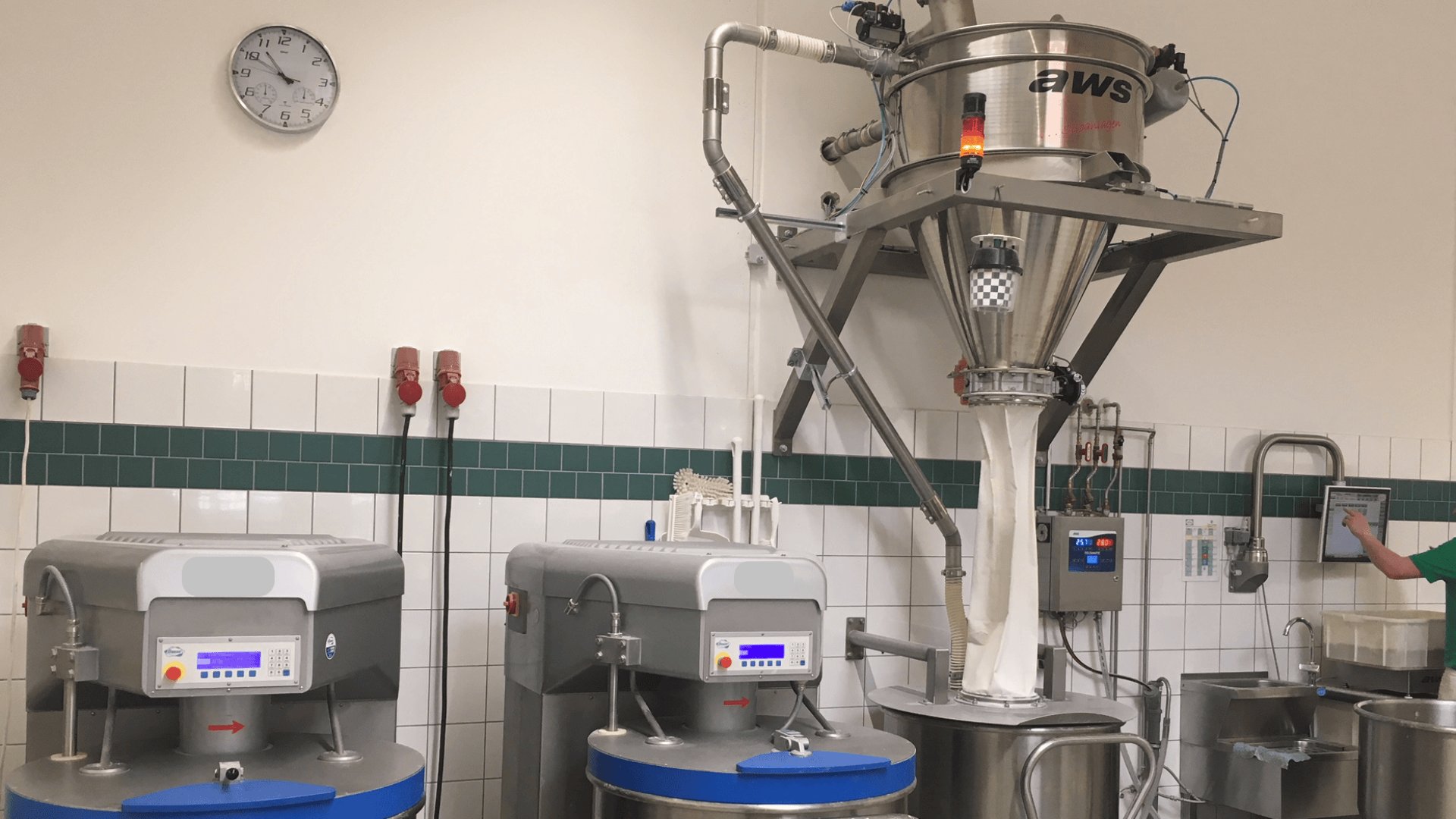 Industrielle Backstube mit großen Mischern und einer Mehlsiebmaschine aus Edelstahl. An der Wand ist eine Uhr angebracht, und auf der rechten Seite des Bildes ist die Hand einer Person zu sehen, die ein Bedienfeld bedient.