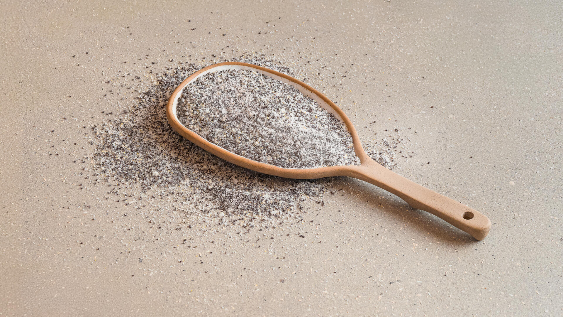 Auf einer hellen Arbeitsplatte liegt ein Holzlöffel, gefüllt mit einer Mischung aus Mohnsamen und Kristallzucker. Um ihn herum ist etwas von der Mischung verschüttet.