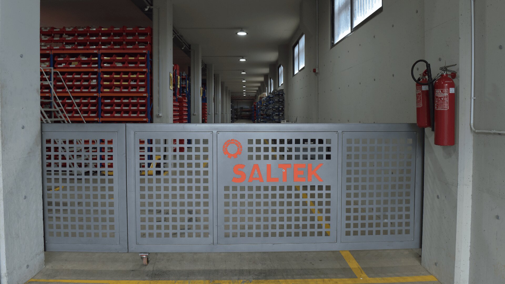 A metal gate with SALTEK written on it blocks the entrance to an industrial storage area filled with shelves and bins. Two red fire extinguishers are mounted on the right wall near the gate.