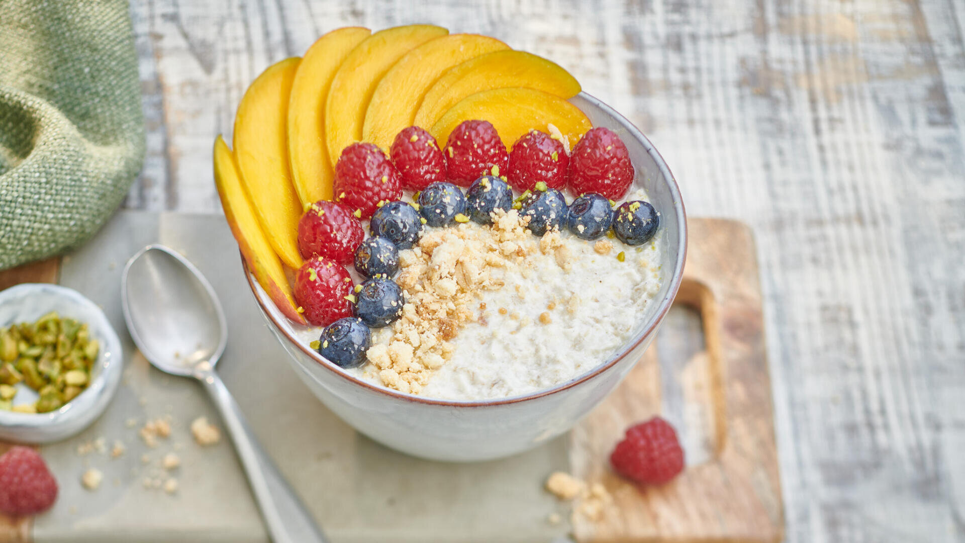 Eine Schüssel Haferbrei mit Mangoscheiben, Himbeeren, Blaubeeren und zerstoßenen Nüssen steht auf einem Holzbrett. Daneben stehen ein Löffel und eine kleine Schale mit Pistazien.