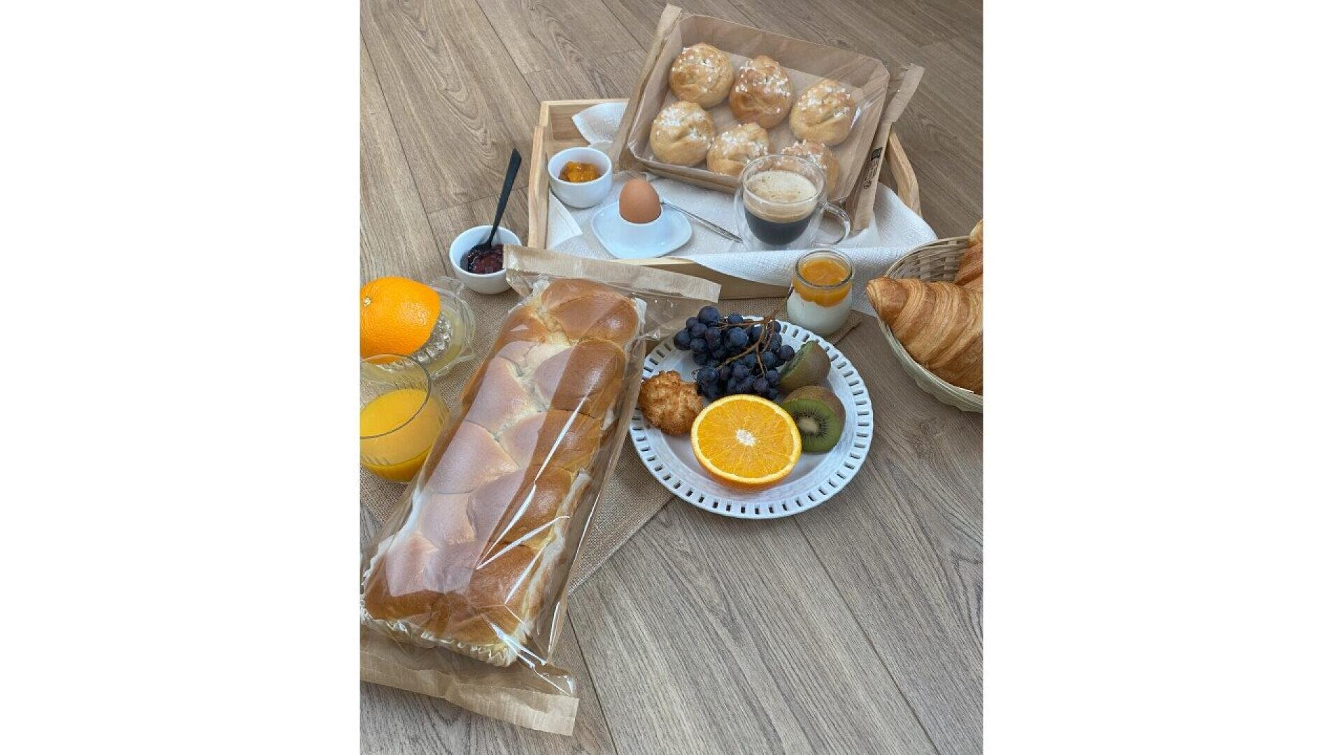 Das Frühstück auf dem Holzboden besteht aus Brötchen, einem Croissant, einer Orange, Weintrauben, Kiwi, einer halben Orange, Saft, einem Ei, Gebäck, Marmelade und Kaffee, alles fein säuberlich auf Tellern und in Körben angeordnet.