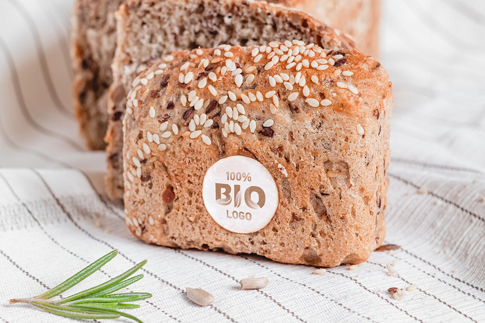Nahaufnahme eines Mehrkornbrotes mit Sesam auf der Oberseite und einem runden Etikett mit der Aufschrift 100% BIO LOGO. Das Brot liegt auf einem weißen Tuch mit braunen Streifen, daneben steht ein Rosmarinzweig.