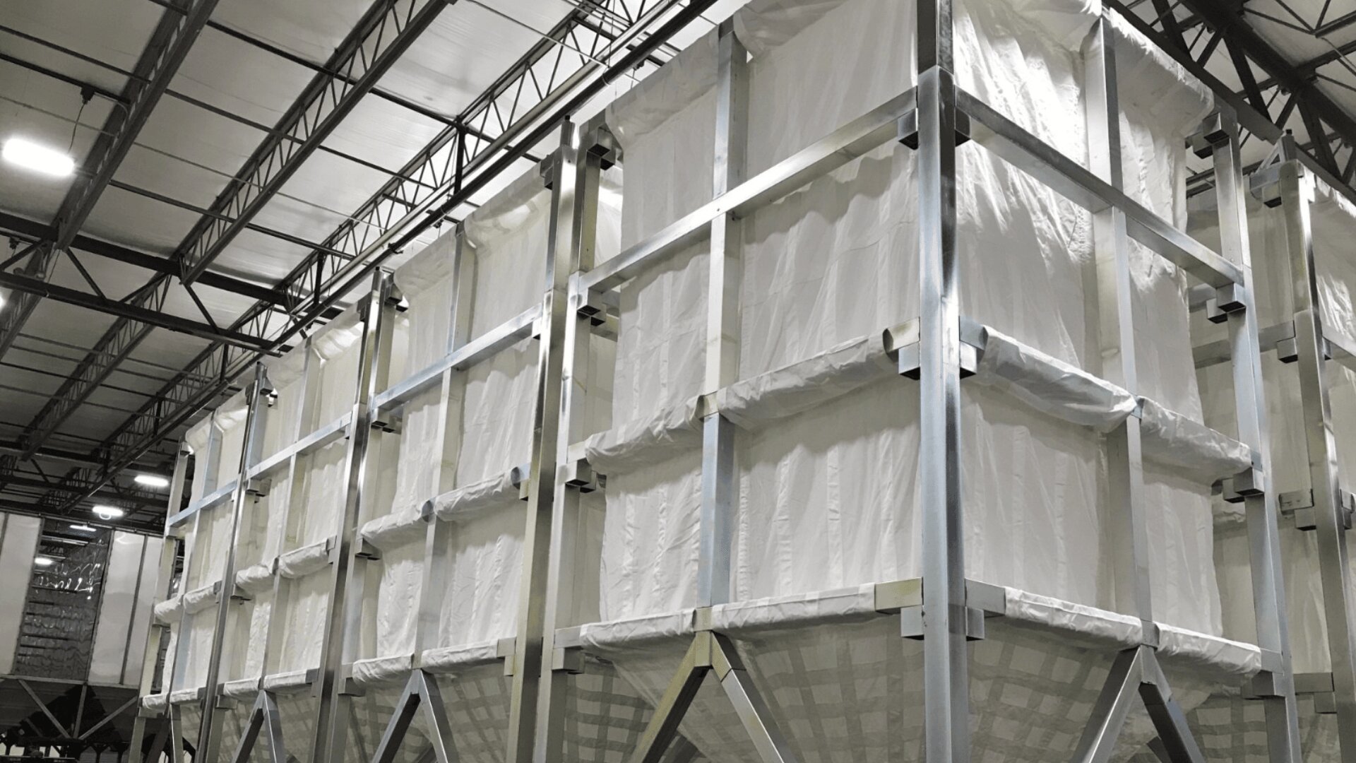 Large white industrial fabric storage bins with metal frames are stacked in rows inside a warehouse with a high ceiling and exposed beams.