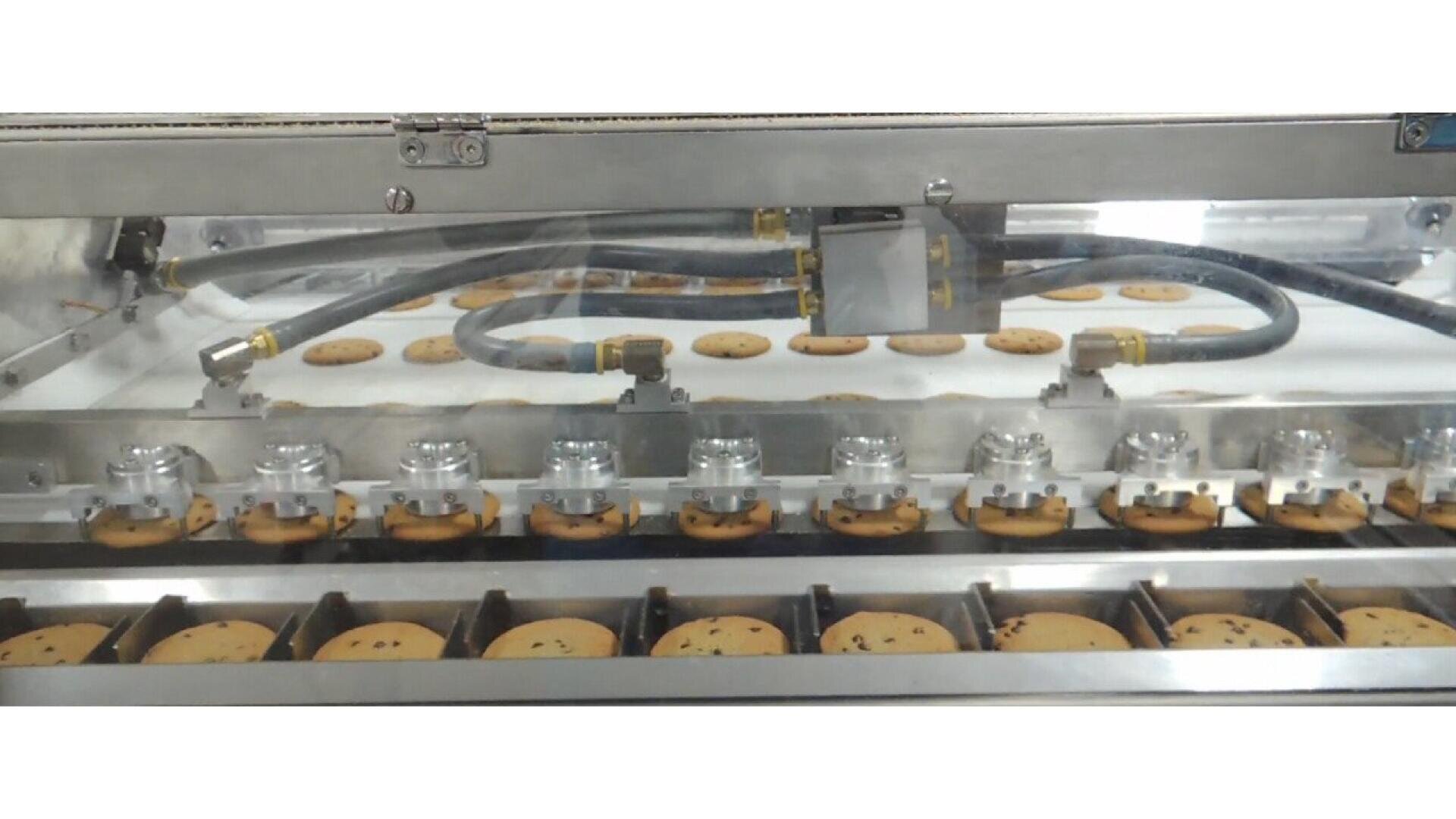 Eine Maschine in einer Fabrik sortiert oder verpackt automatisch Reihen von Schokokeksen auf einem Förderband, wobei Metall- und Kunststoffteile sichtbar sind.