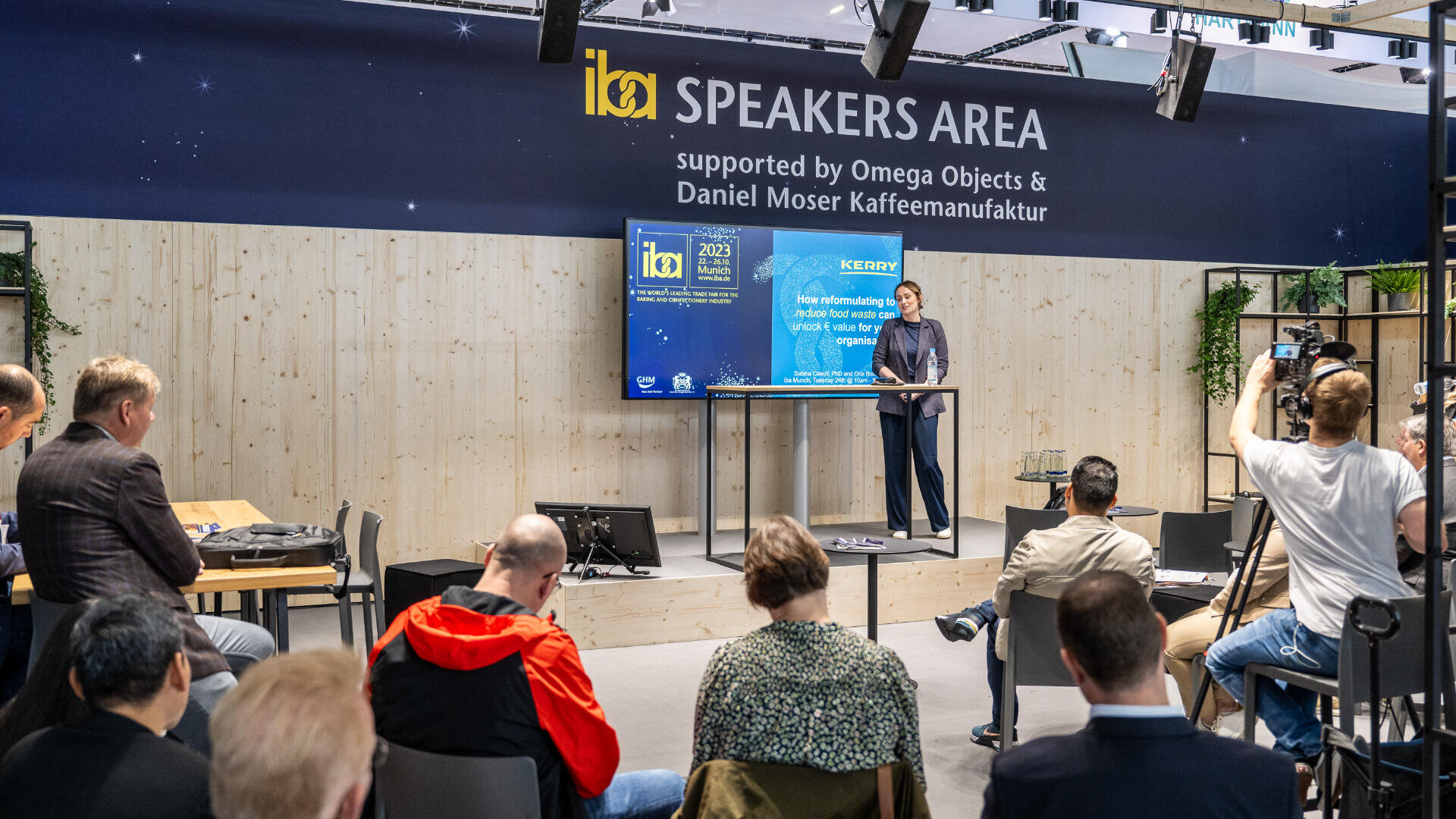 Ein Redner steht an einem Podium und hält einen Vortrag vor einem Publikum in einem Konferenzbereich mit der Bezeichnung „Speakers Area“. Kameras zeichnen auf, und hinter ihm befindet sich eine große Präsentationsleinwand. Das Publikum sitzt da und hört aufmerksam zu.