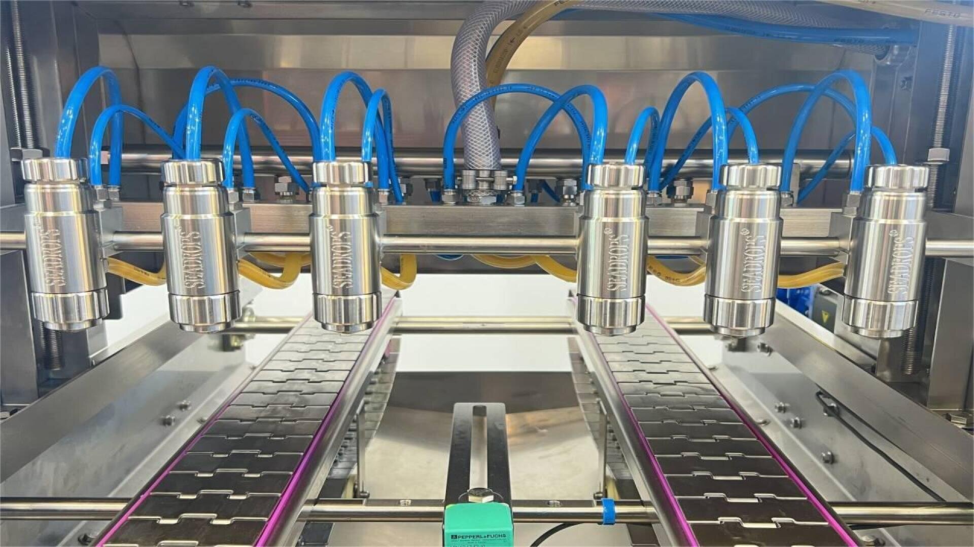 Automated liquid filling machine with six metal nozzles and blue hoses positioned above a conveyor belt, ready to dispense liquid into containers in an industrial setting.