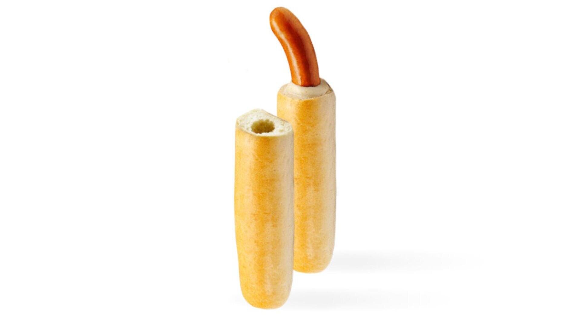 Eine Hotdog-Wurst, die teilweise in ein ausgehöhltes Brötchen gesteckt ist, daneben steht ein weiteres leeres Brötchen, beides vor einem weißen Hintergrund.