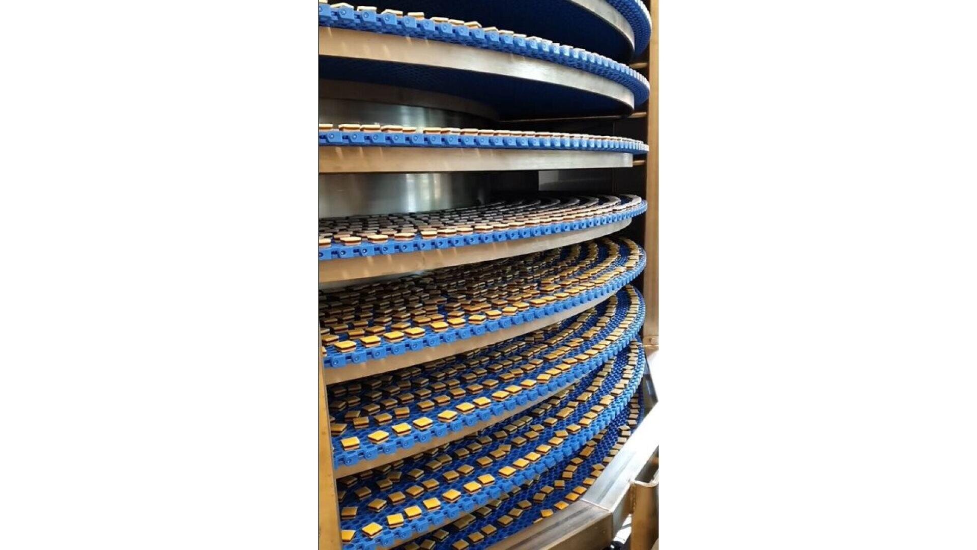 A spiral conveyor belt system with multiple blue tiers, each carrying rows of small, rectangular yellow food items, likely moving upward through the machine.