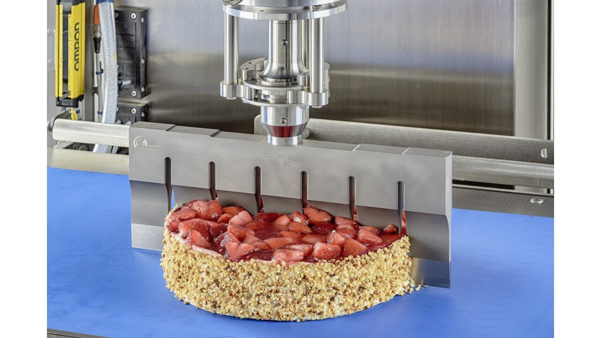 A machine with a multi-blade cutter is slicing a round strawberry cake topped with fruit and nuts on the sides, placed on a blue surface.