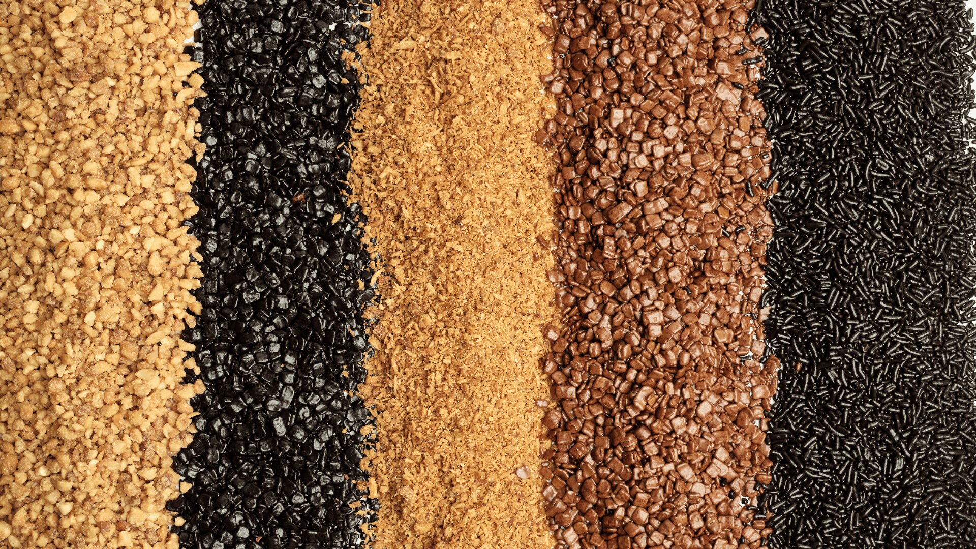 Five vertical stripes of different grains or seeds in colors from left to right: light yellow, black, golden brown, reddish brown, and dark brown, arranged side by side in textured rows.