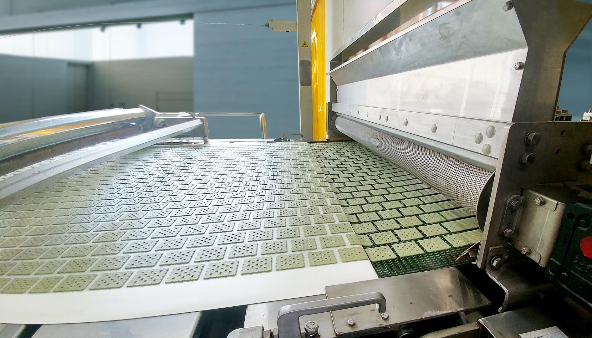 A food production machine shapes and cuts uniform square crackers, which move along a conveyor belt in a bright, industrial facility.