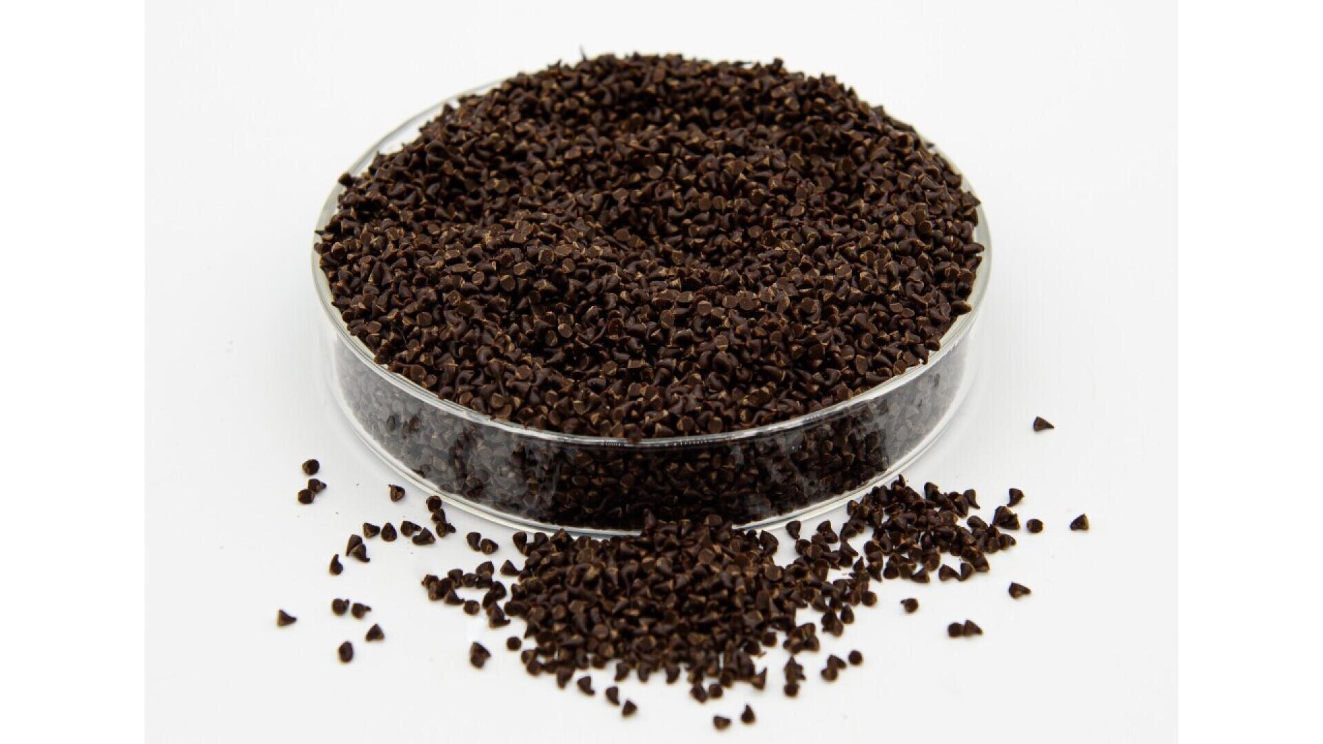 Eine durchsichtige, runde Petrischale, gefüllt mit kleinen, dunkelbraunen Körnchen, steht auf einer weißen Oberfläche, wobei einige Körnchen über die Schale verstreut sind.