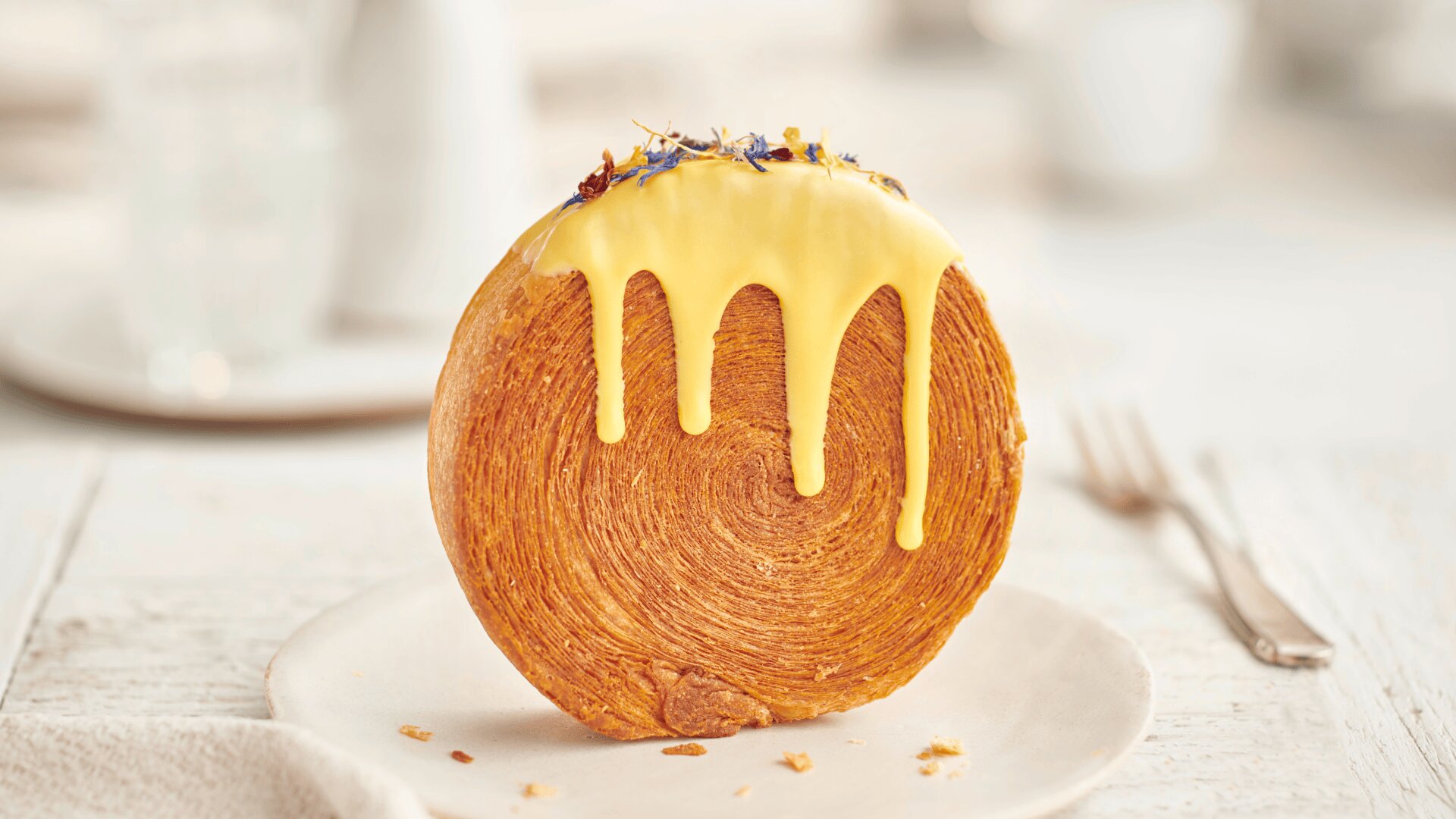 Ein rundes, geschichtetes Gebäck steht aufrecht auf einem Teller und ist mit gelber Glasur überzogen, die an den Seiten heruntertropft. Der Hintergrund ist leicht verschwommen, eine Gabel und Geschirr sind zu sehen.