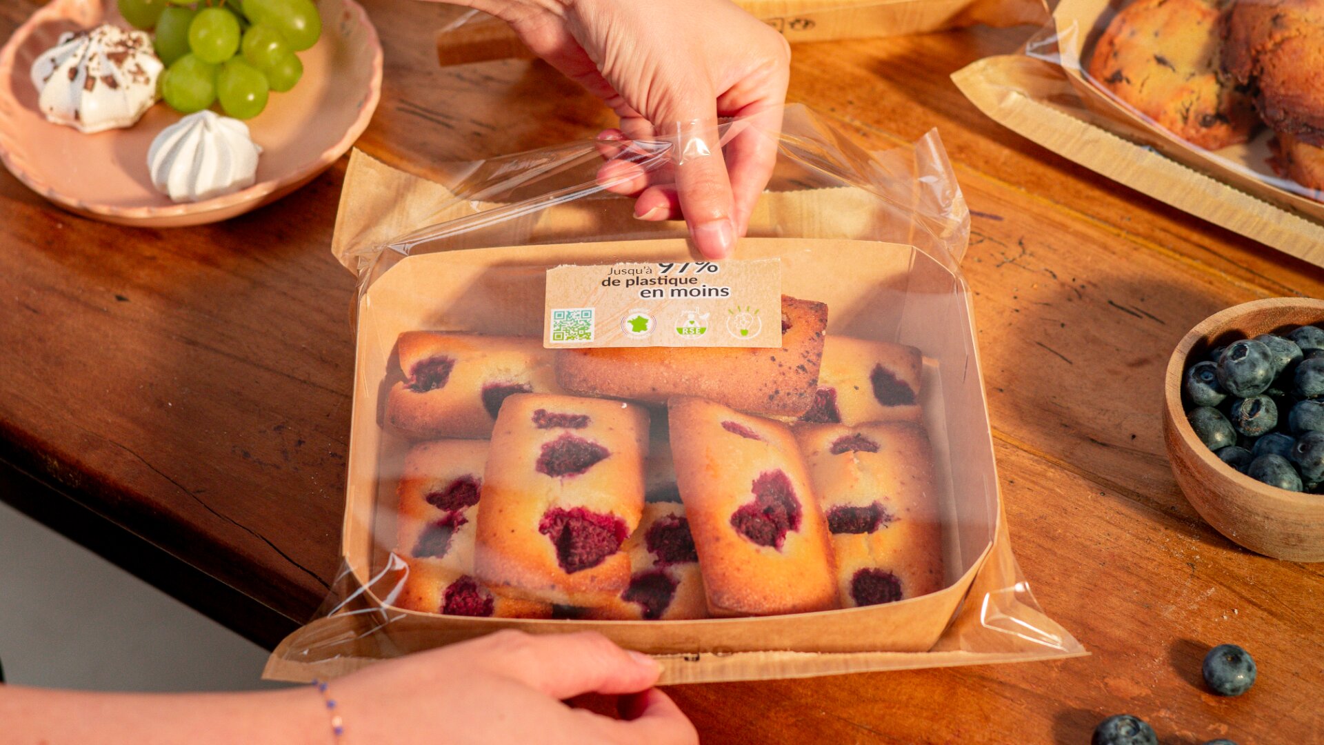 A person seals a cardboard tray of rectangular raspberry pastries with clear plastic wrap. The tray sits on a wooden table next to a plate of green grapes and meringues and a bowl of blueberries.
