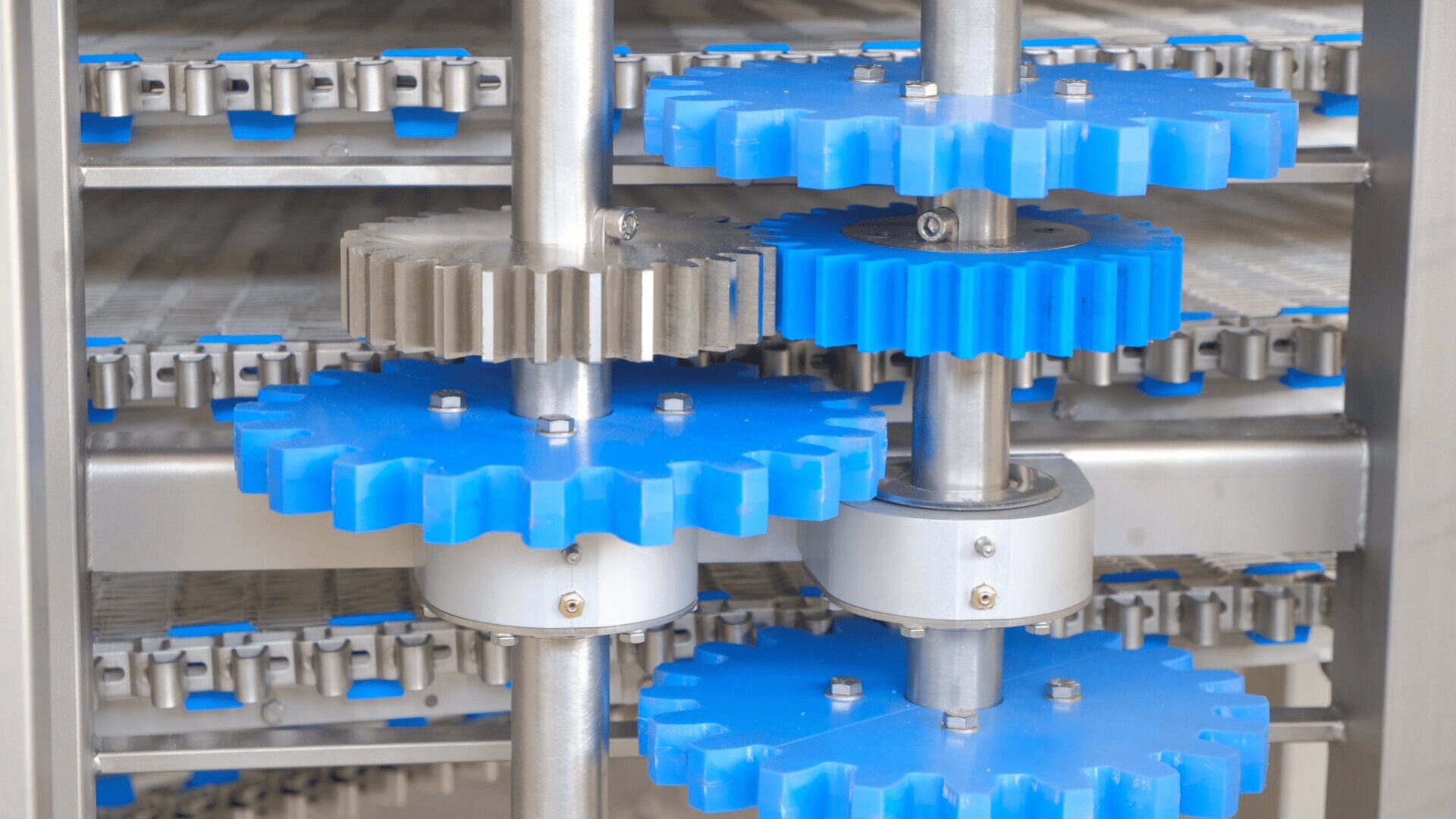 Close-up view of several large blue and silver gears in a mechanical system, possibly part of industrial machinery or equipment, with metal rods and conveyor belts visible in the background.
