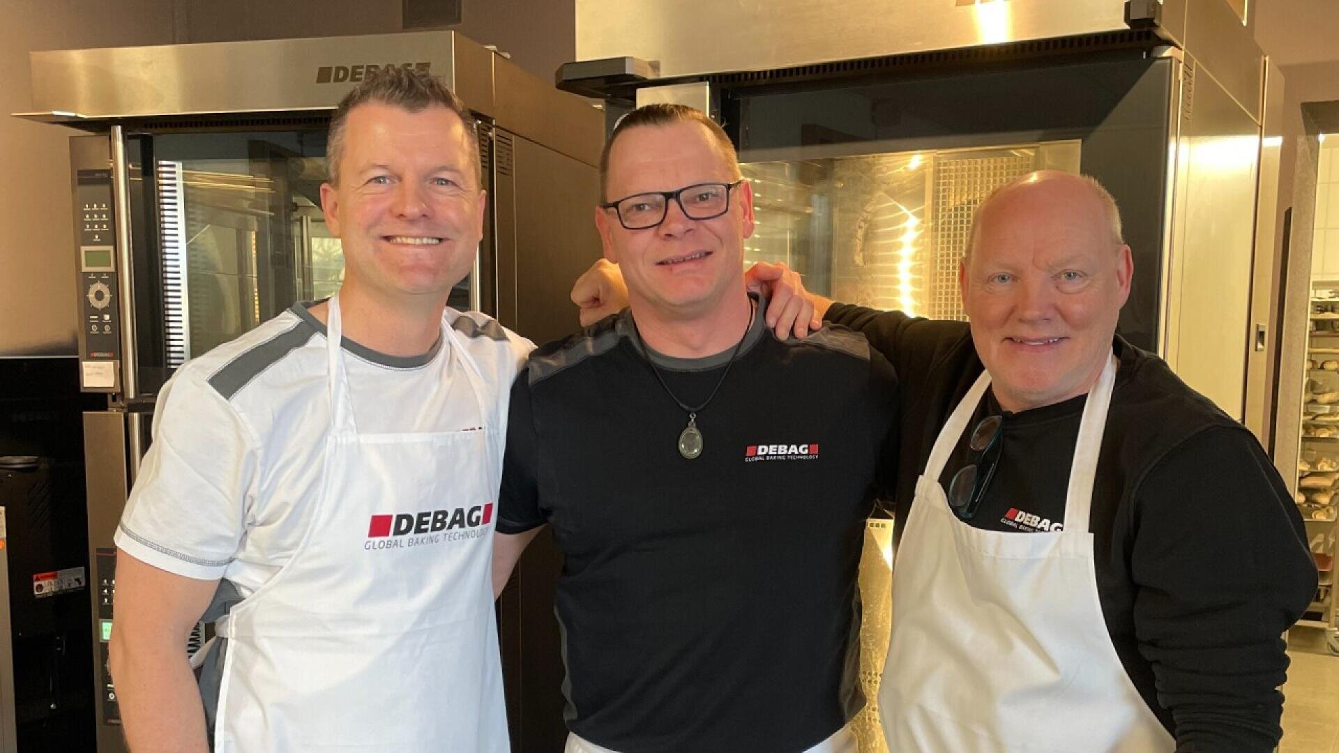 Three men stand together smiling in a commercial kitchen, all wearing Debag-branded shirts and two wearing white aprons. Industrial ovens are visible in the background.