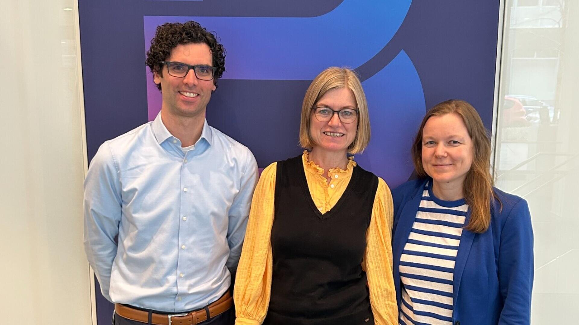 Three people stand indoors in front of a blue and purple backdrop, smiling at the camera. The man on the left wears glasses and a light blue shirt. The two women wear yellow and blue tops.