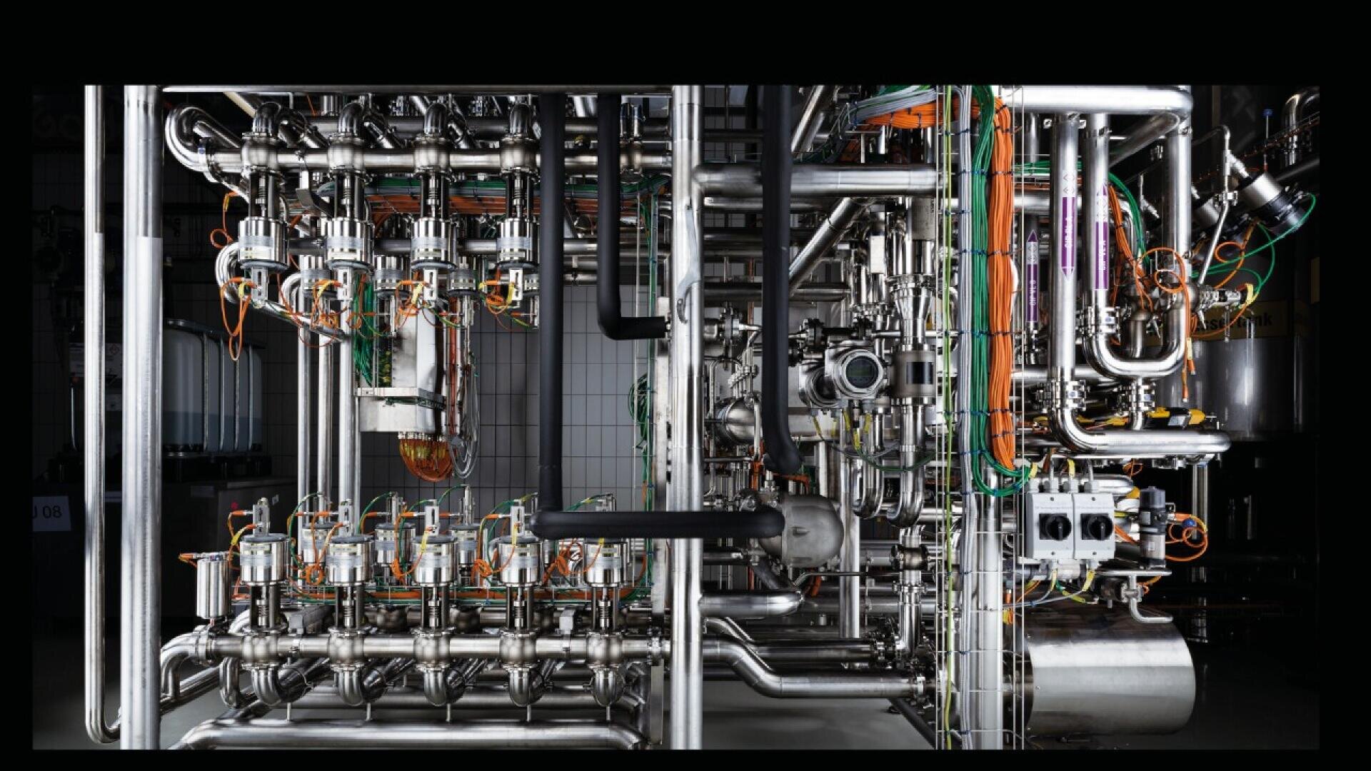 Complex industrial machinery with numerous stainless steel pipes, valves, gauges, and orange and green electrical wires, set against a tiled wall background.
