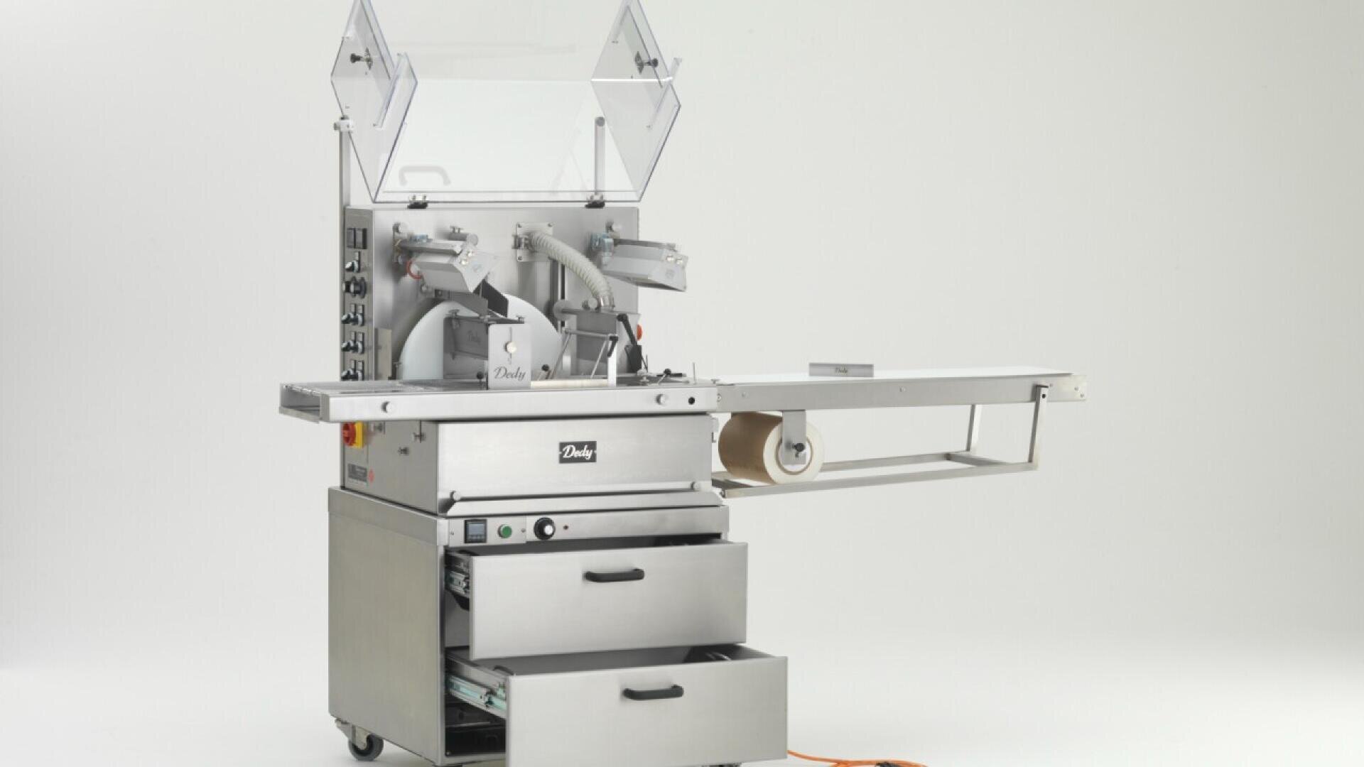 A stainless steel commercial food processing machine with an attached conveyor belt, control panels, two drawers, and a clear protective cover, standing on wheels in a white studio setting.
