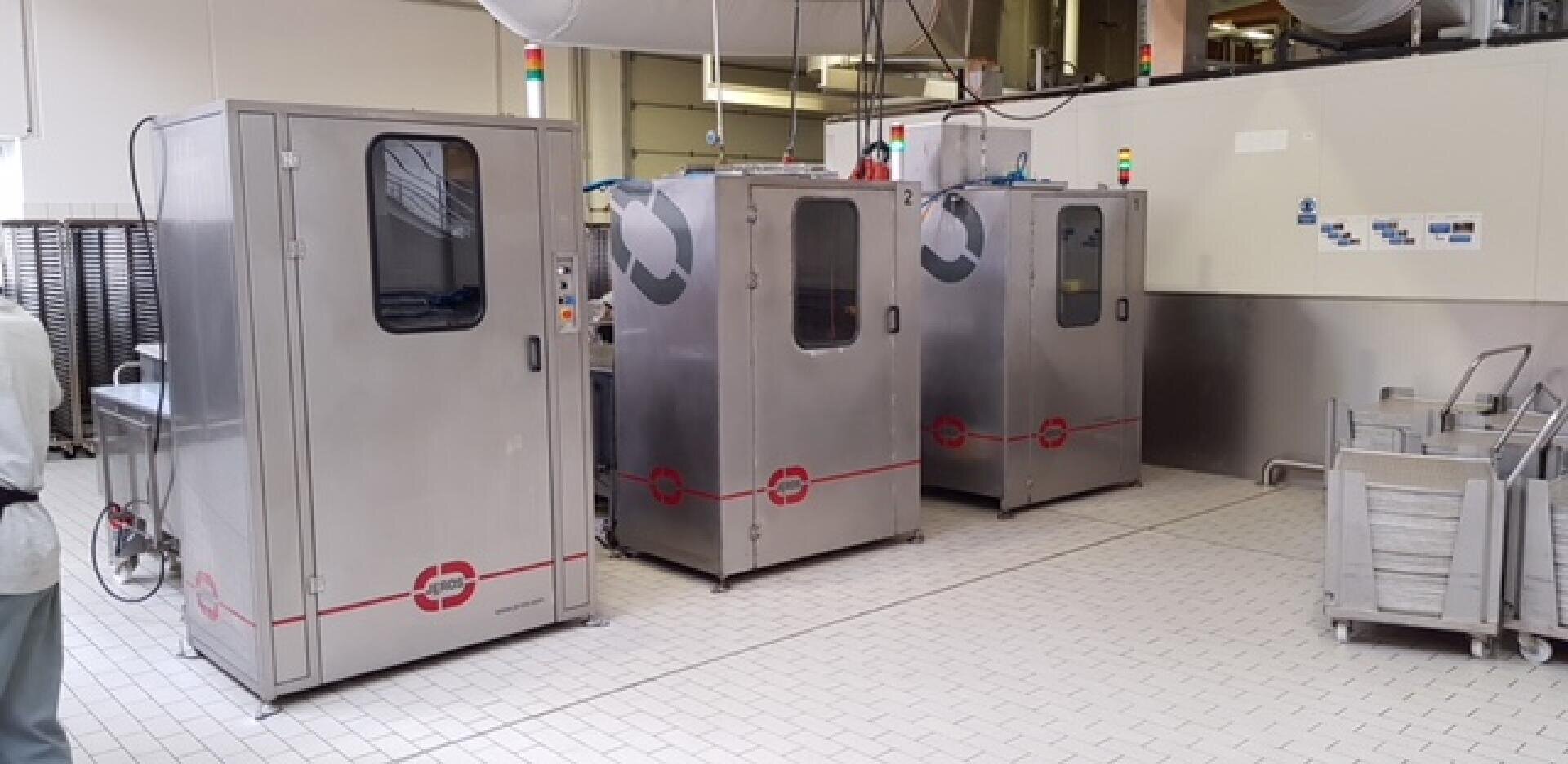 Three large stainless steel industrial machines with glass doors stand in a clean, tiled facility. Stacks of trays and carts are nearby, and overhead pipes run along the ceiling.