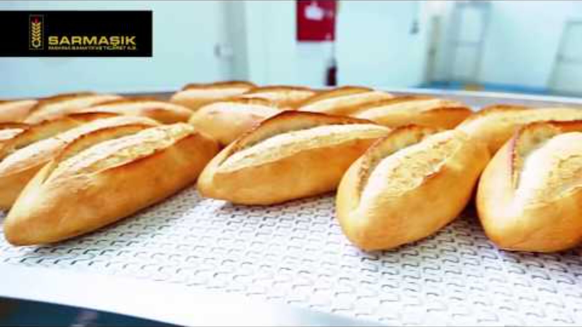 Several freshly baked loaves of bread are neatly arranged on a conveyor belt in a bakery, with the Sarmasik logo visible in the top left corner of the image.