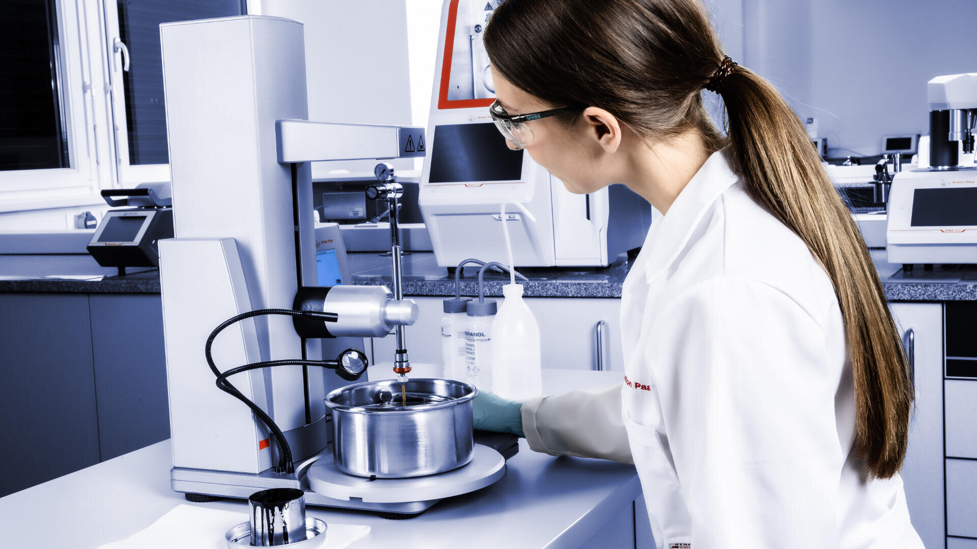 Eine Frau in einem Laborkittel und mit Schutzbrille bedient wissenschaftliche Geräte in einem Labor und überwacht einen Metallbehälter auf einer Maschine. Das Labor ist hell und modern, mit verschiedenen Instrumenten und einer Flasche in der Nähe.