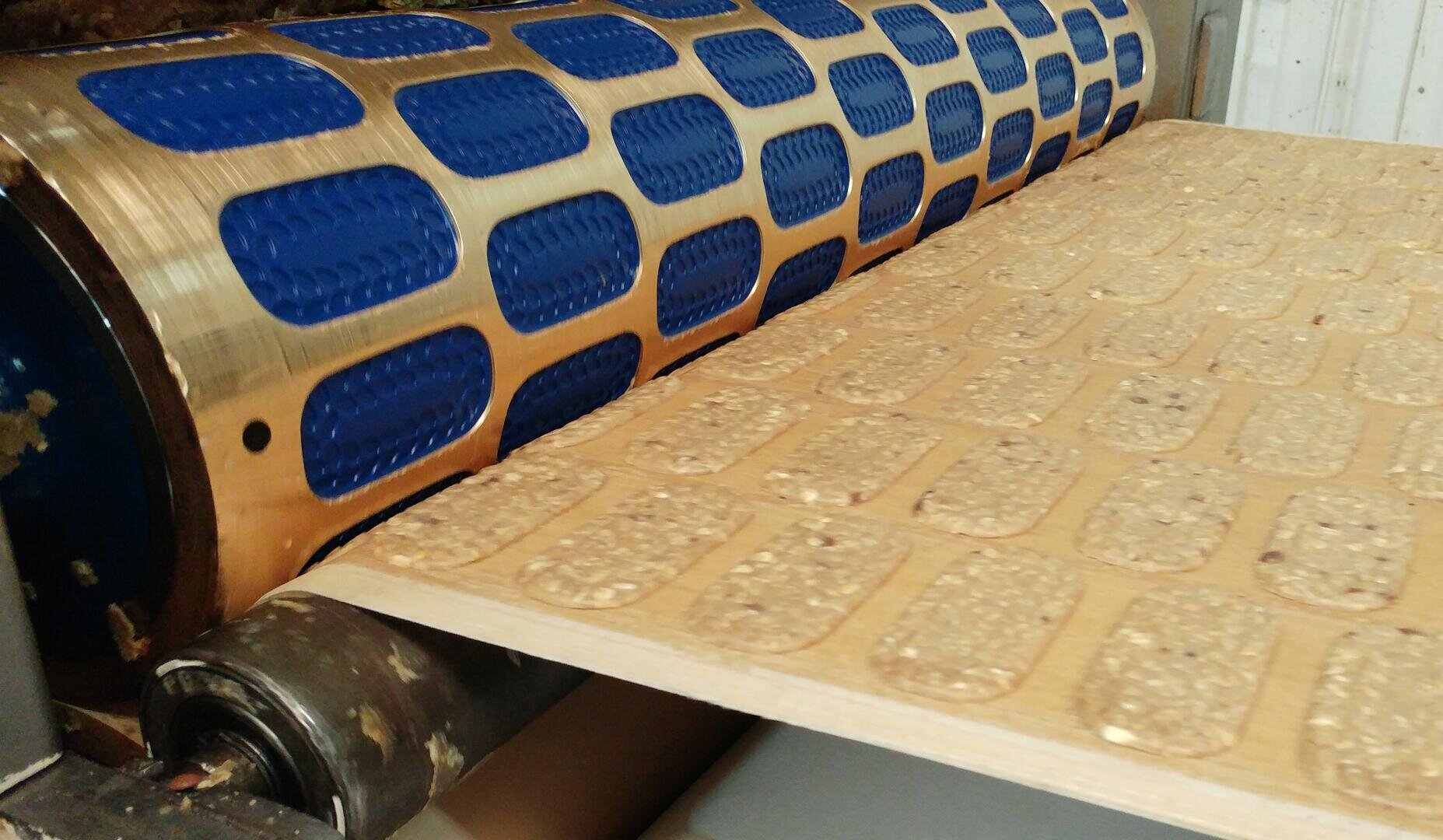 A close-up of a machine pressing and shaping dough into uniform rectangular cracker shapes with textured surfaces on a conveyor belt.