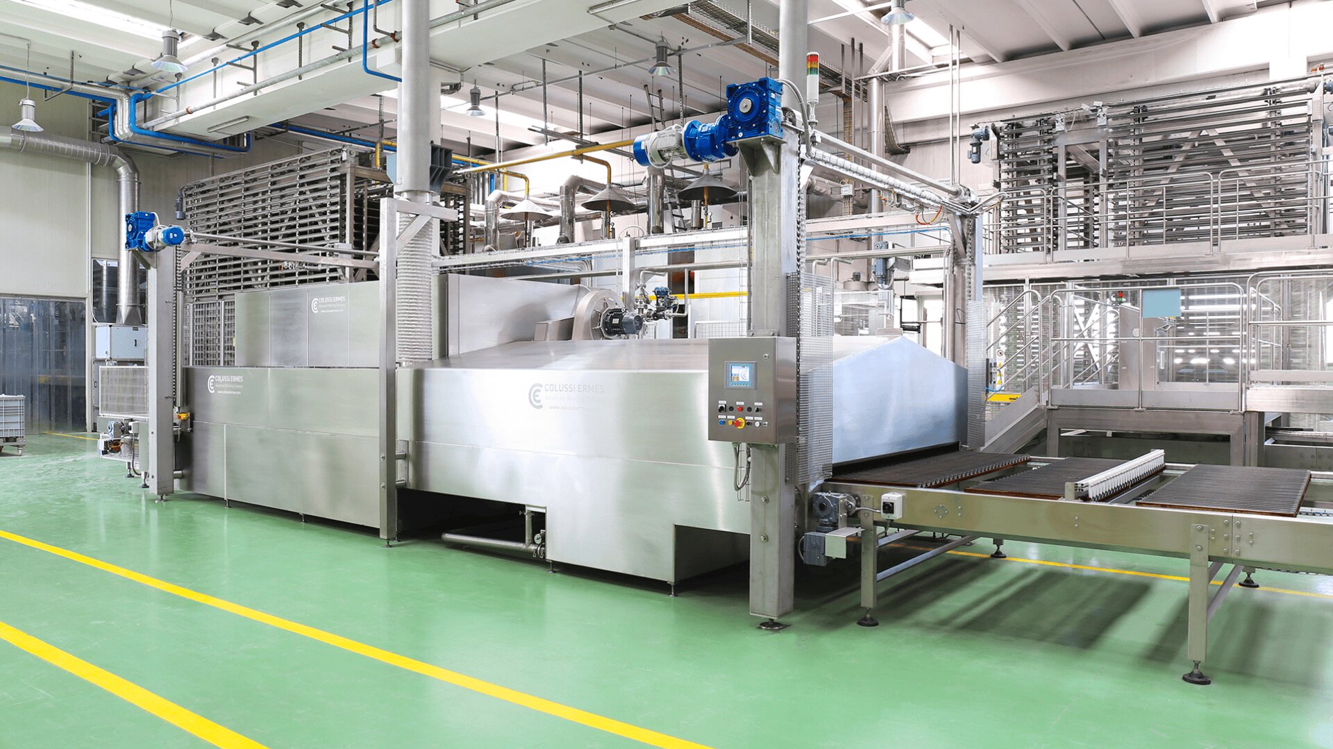 Große industrielle Lebensmittelverarbeitungsmaschine aus rostfreiem Stahl mit Förderbändern in einer sauberen Fabrikumgebung, mit Schalttafeln und Überkopfverrohrung, auf grünem Boden mit gelben Sicherheitslinien.