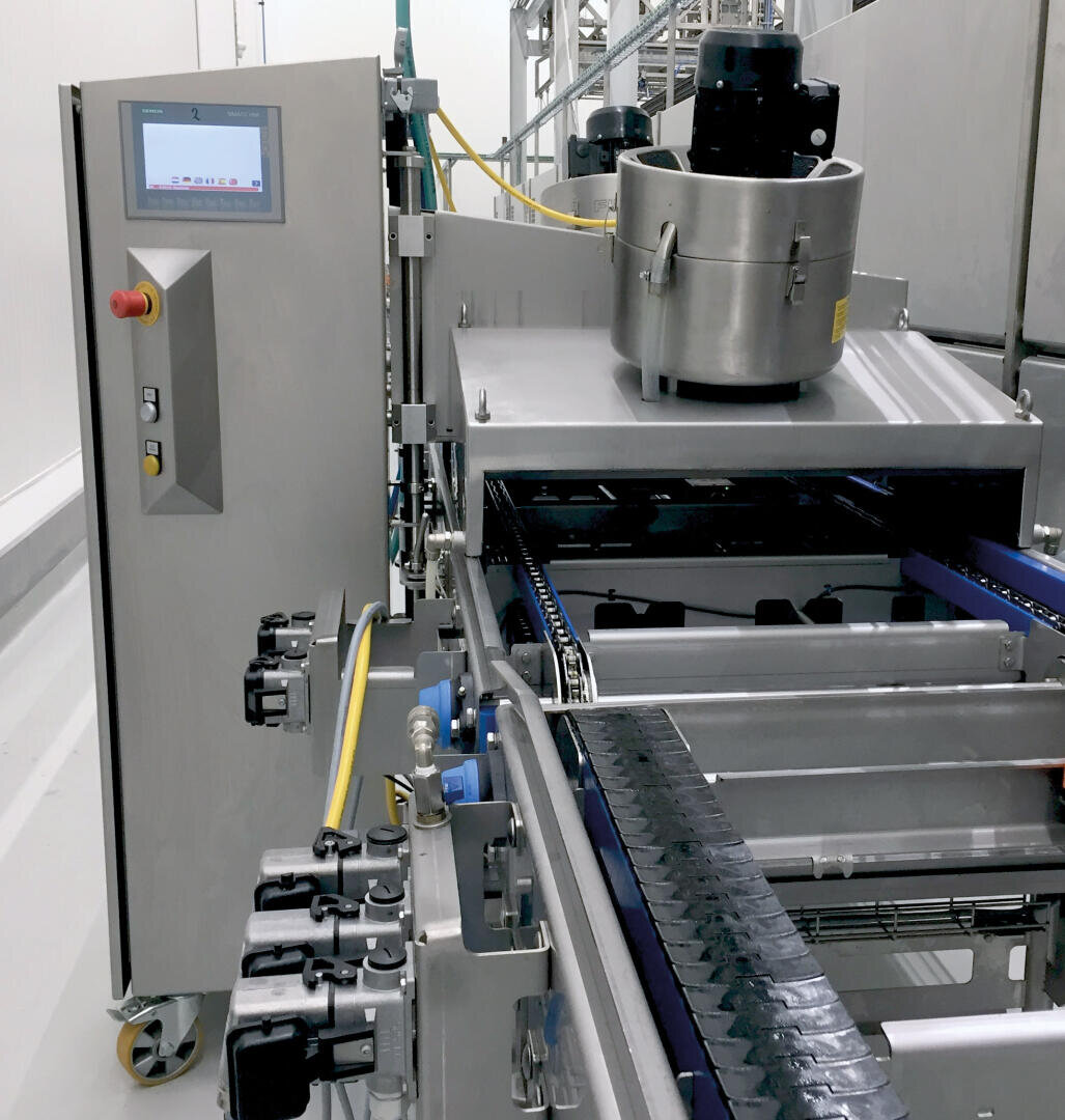 A stainless steel industrial machine with a control panel and touchscreen display, featuring conveyor belts and a large metal hopper, located in a clean, modern facility.