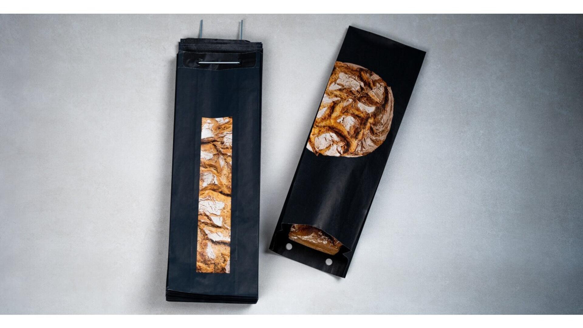 Two black paper bread bags with clear windows displaying rustic artisan loaves inside, placed on a light gray surface. One bag is upright and closed, the other is lying down and partially open.