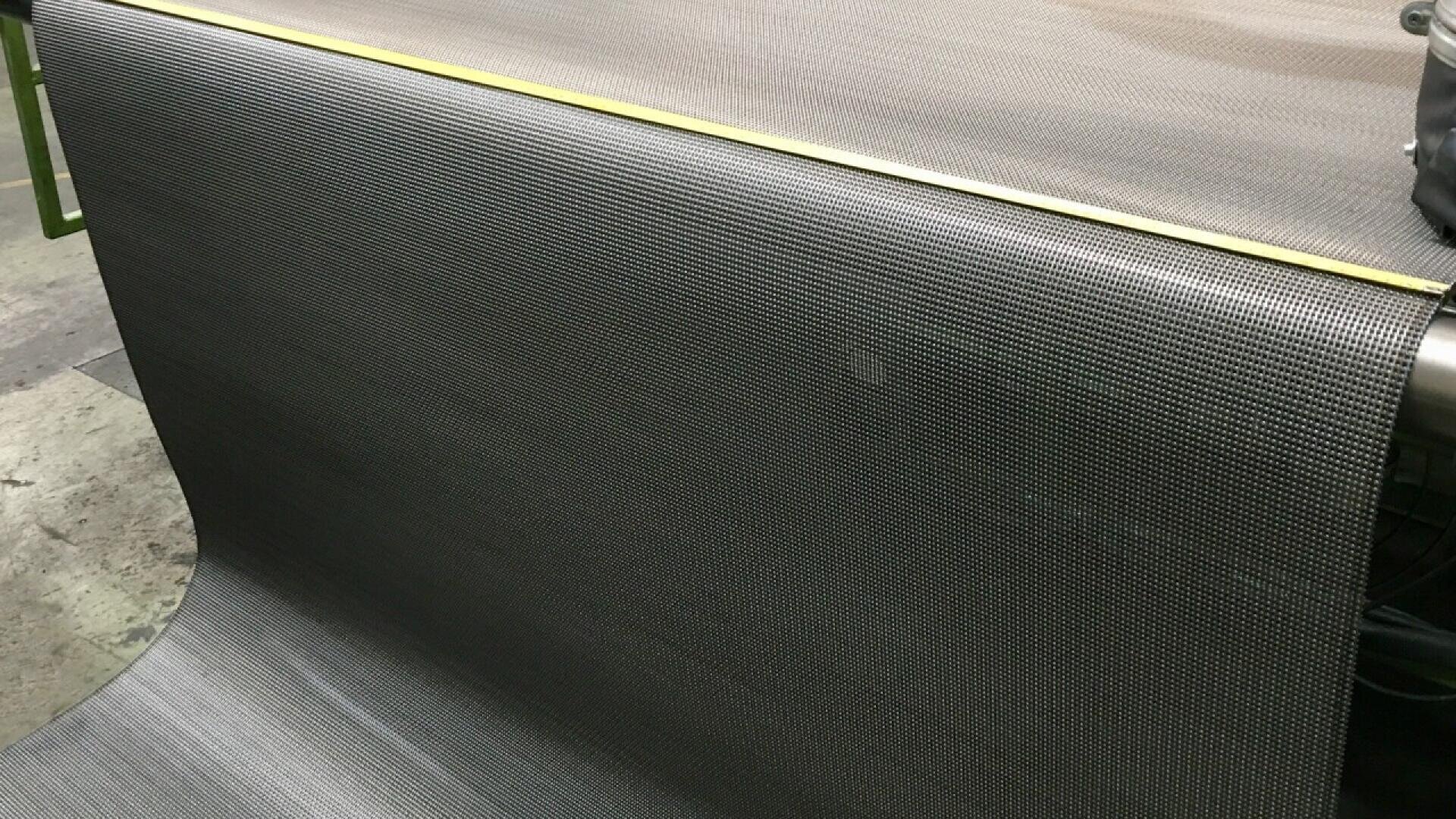 A large roll of fine metal mesh fabric is being unwound onto a flat industrial surface, with a yellow measuring tape running along the edge.