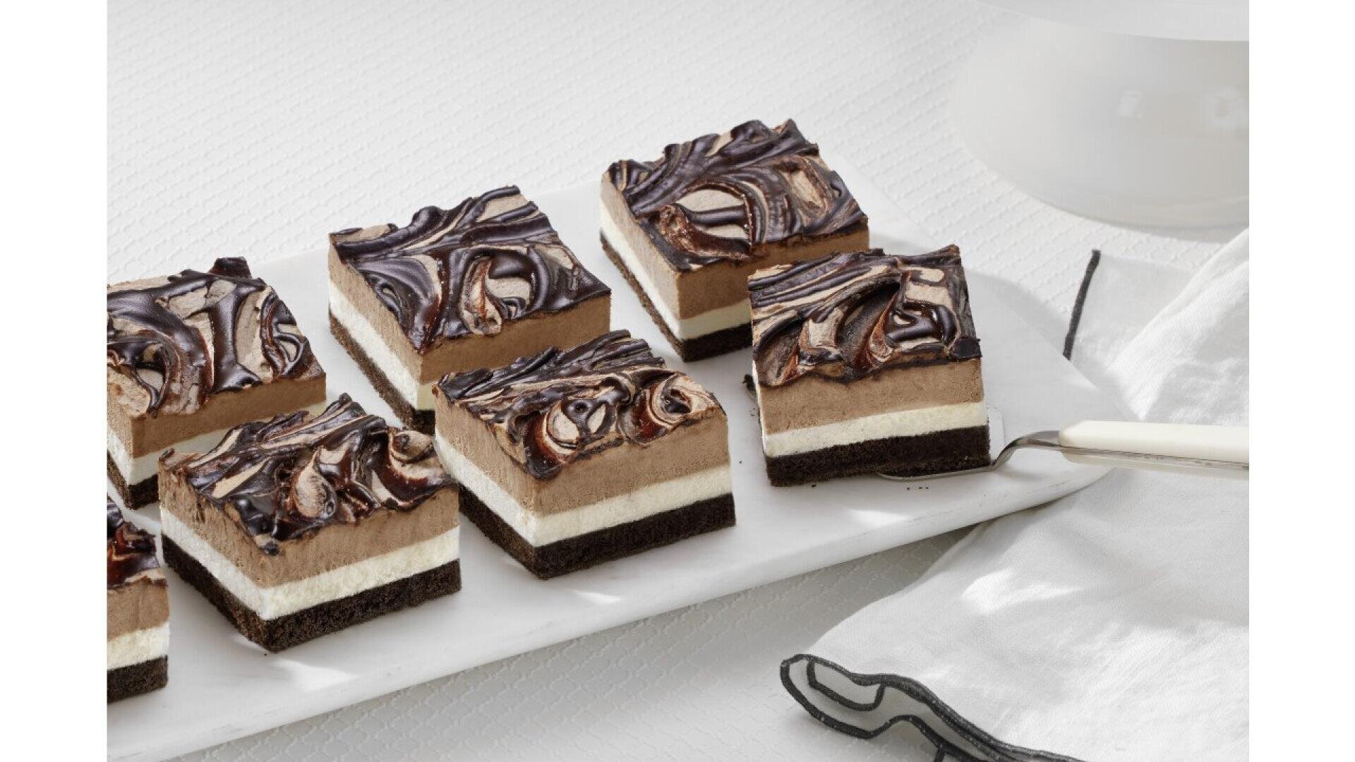 Nine layered dessert bars with a chocolate base, cream and chocolate mousse layers, topped with a swirled chocolate and vanilla design, arranged on a white rectangular tray beside a napkin and serving utensil.