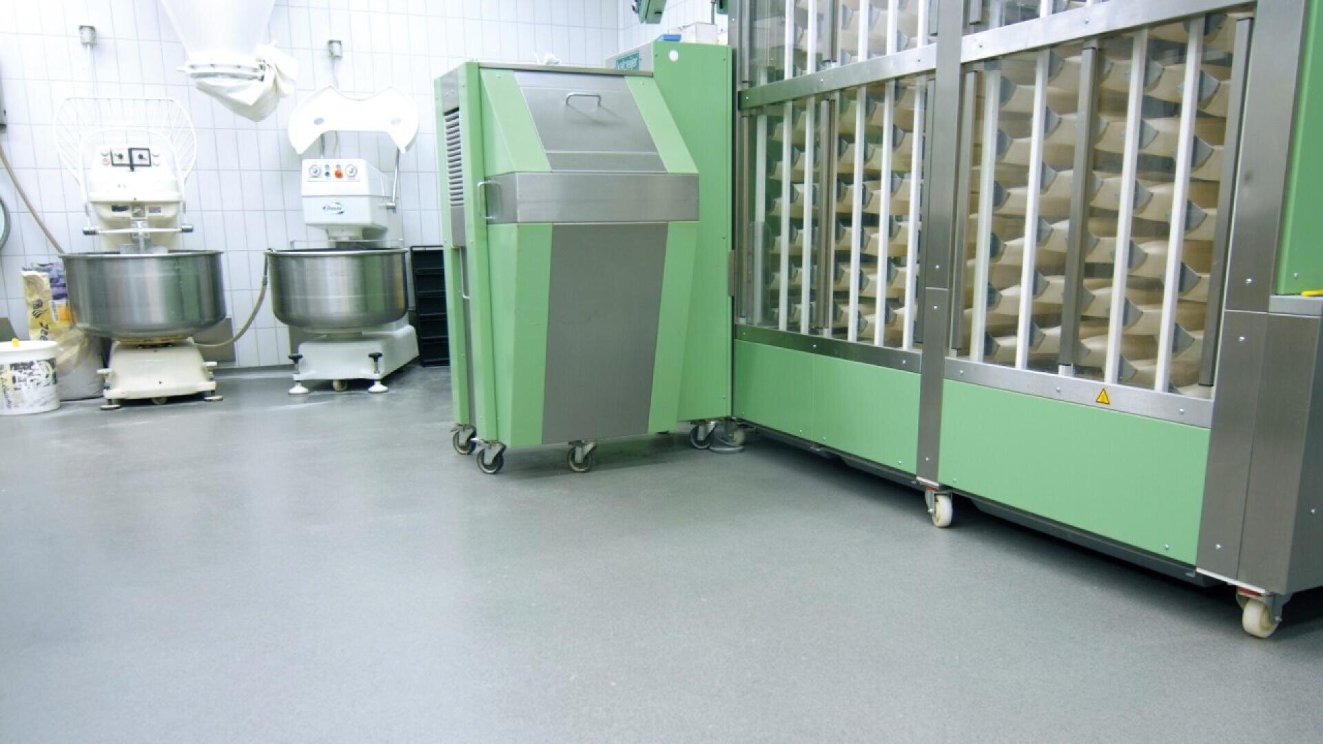 A clean, modern industrial kitchen with large green and silver baking machines, metal mixing bowls, and tiled walls, all arranged on a spotless gray floor.