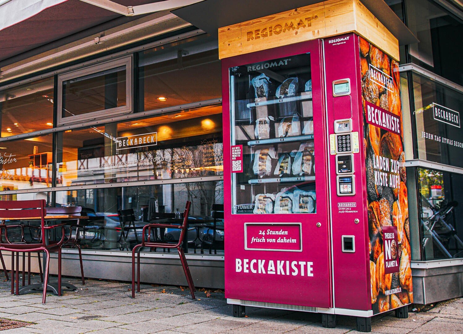 Ein roter Verkaufsautomat mit der Aufschrift Beckakiste steht auf dem Gehweg vor einem Café mit Außenbestuhlung. Der Automat enthält Bäckereiprodukte und Brot, im Hintergrund ist eine moderne Glasfront zu sehen.
