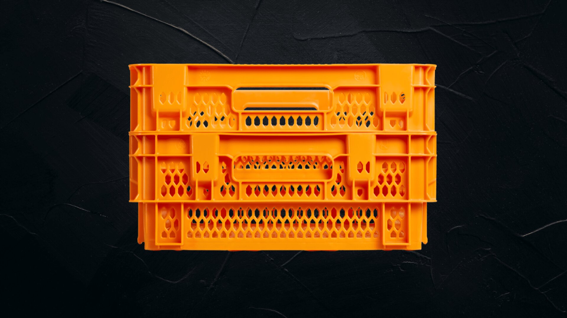Eine leuchtend orangefarbene Kunststoffkiste mit Lochmuster und eingebauten Griffen steht vor einem strukturierten, schwarzen Hintergrund.