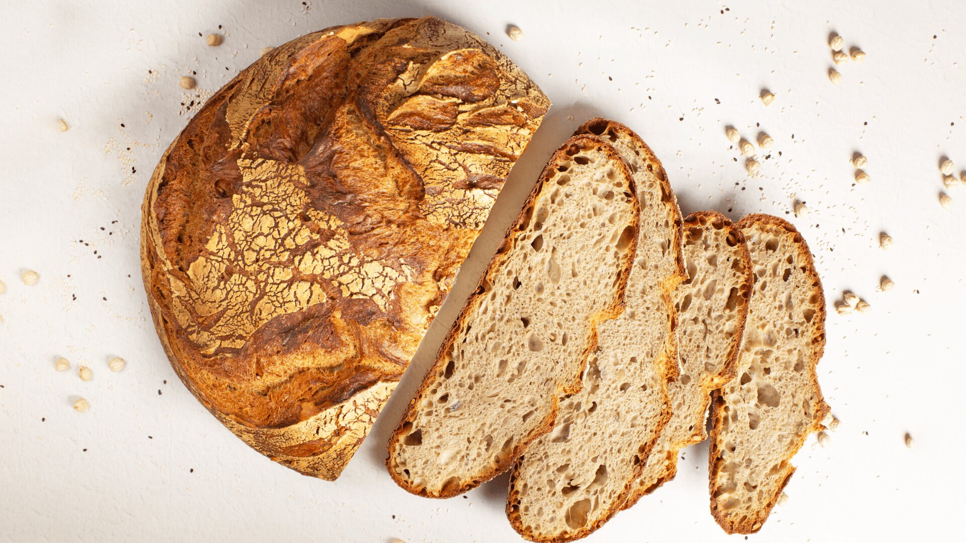 Ein runder Laib rustikalen Brotes, der teilweise aufgeschnitten ist und drei dicke Scheiben auf einer weißen Oberfläche zeigt, um die herum Krümel und Samen verstreut sind.