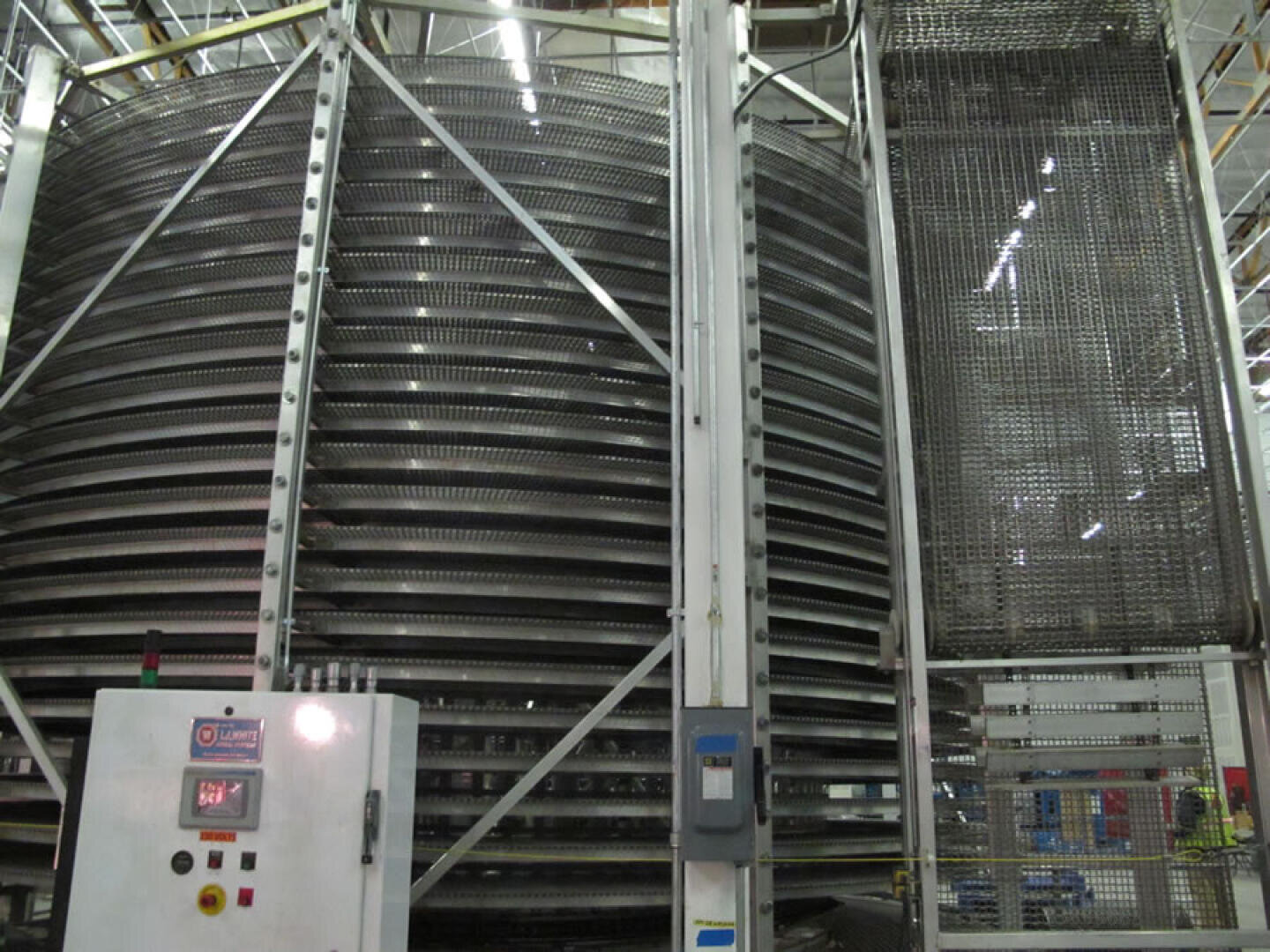 Large industrial spiral conveyor system with multiple stacked metal belts. A control panel with buttons and a display is visible in the foreground. The setting appears to be inside a factory or food processing plant.
