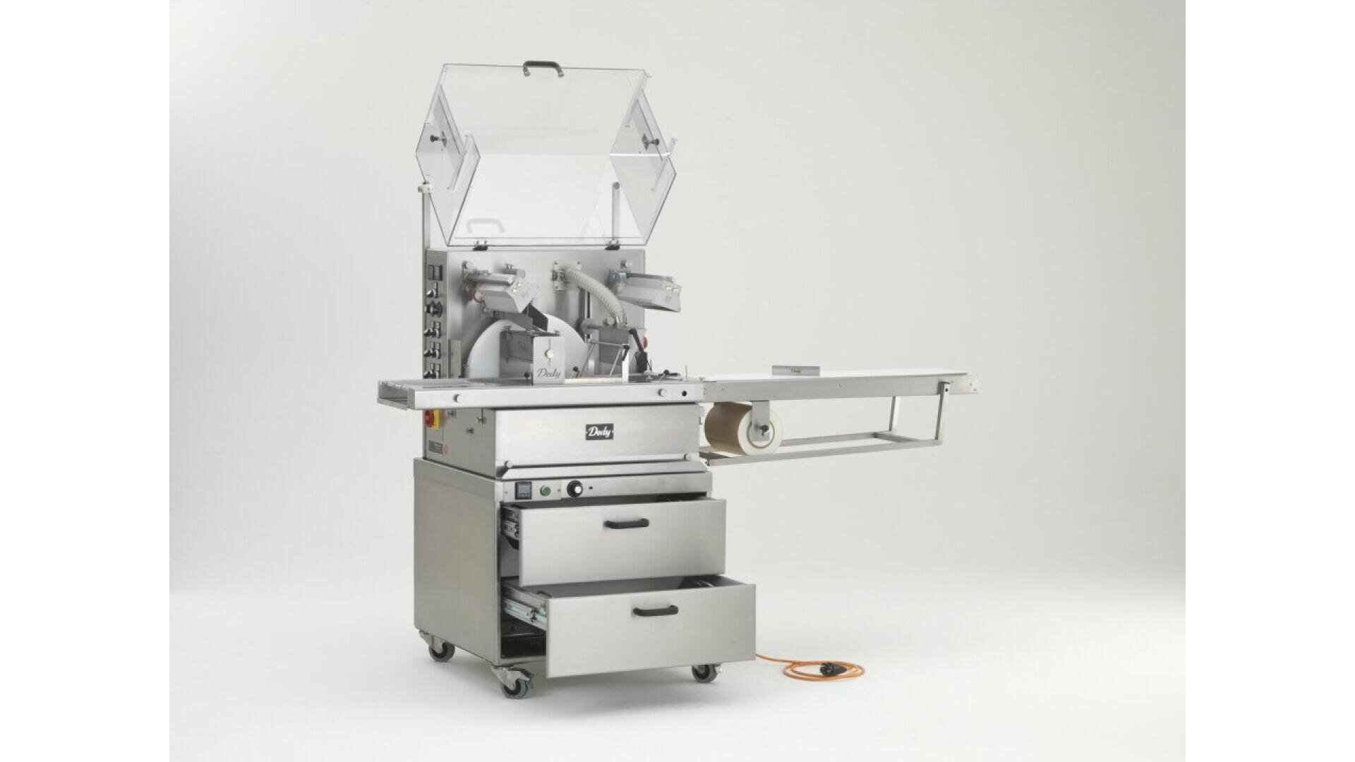 A commercial food packaging machine with a transparent lid, control panel, two drawers, and an extended conveyor tray. The machine is on wheels and has an orange power cord on the floor.